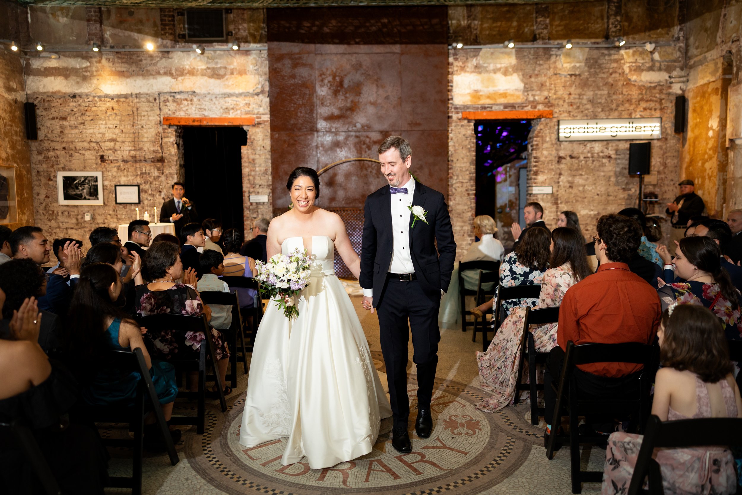 Bride and groom walking down the aisle at a wedding ceremony surrounded by seated guests in a rustic indoor venue with exposed brick walls and warm lighting.