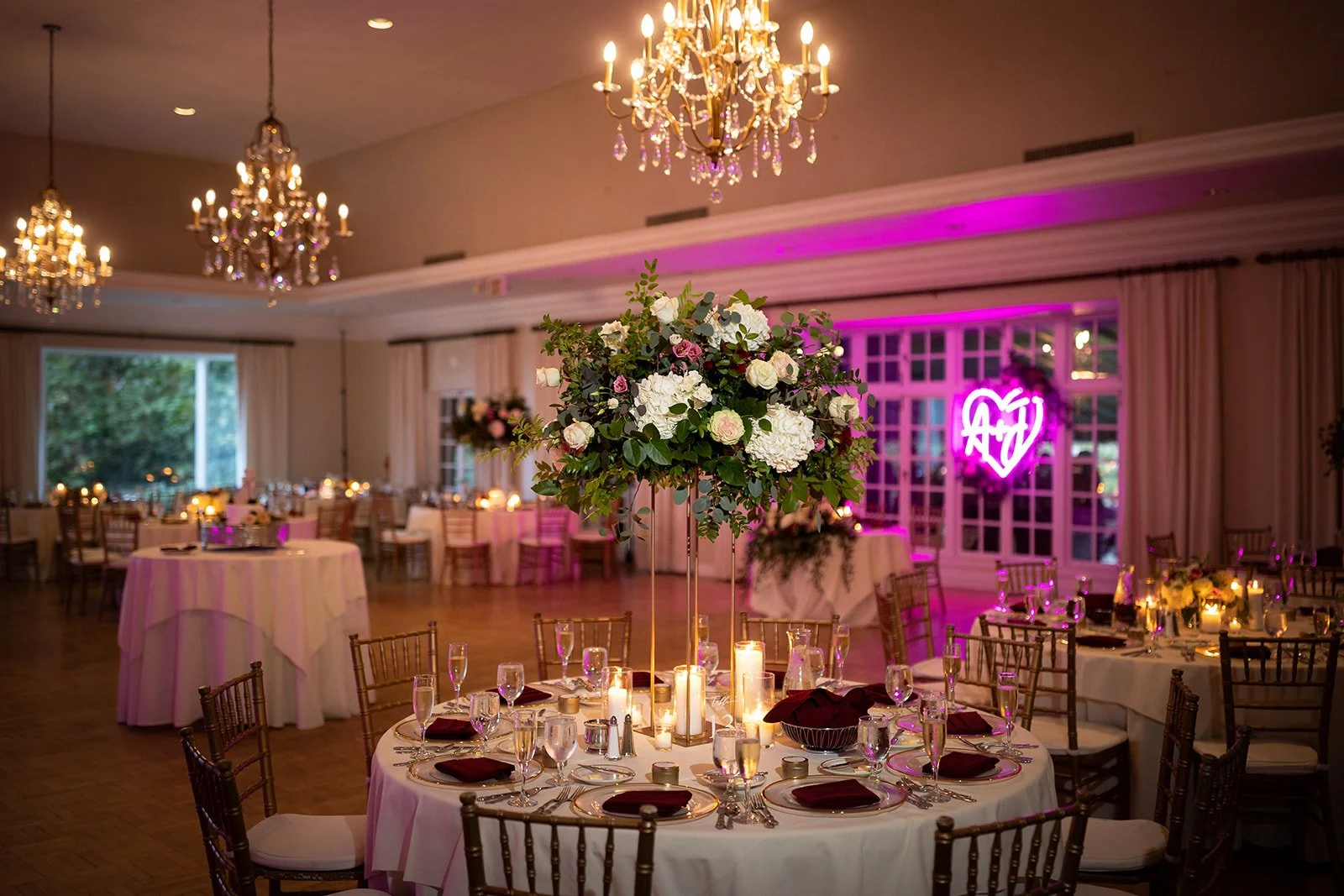 Elegant indoor wedding reception with round tables, tall floral centerpieces, candles, and chandeliers, featuring a neon pink heart with the initials 'AD' in the background.