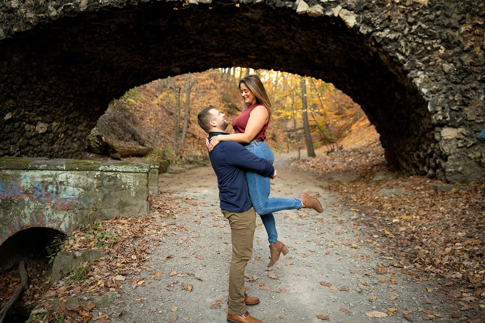 A man lifting a woman under an autumnal stone bridge in a wooded area