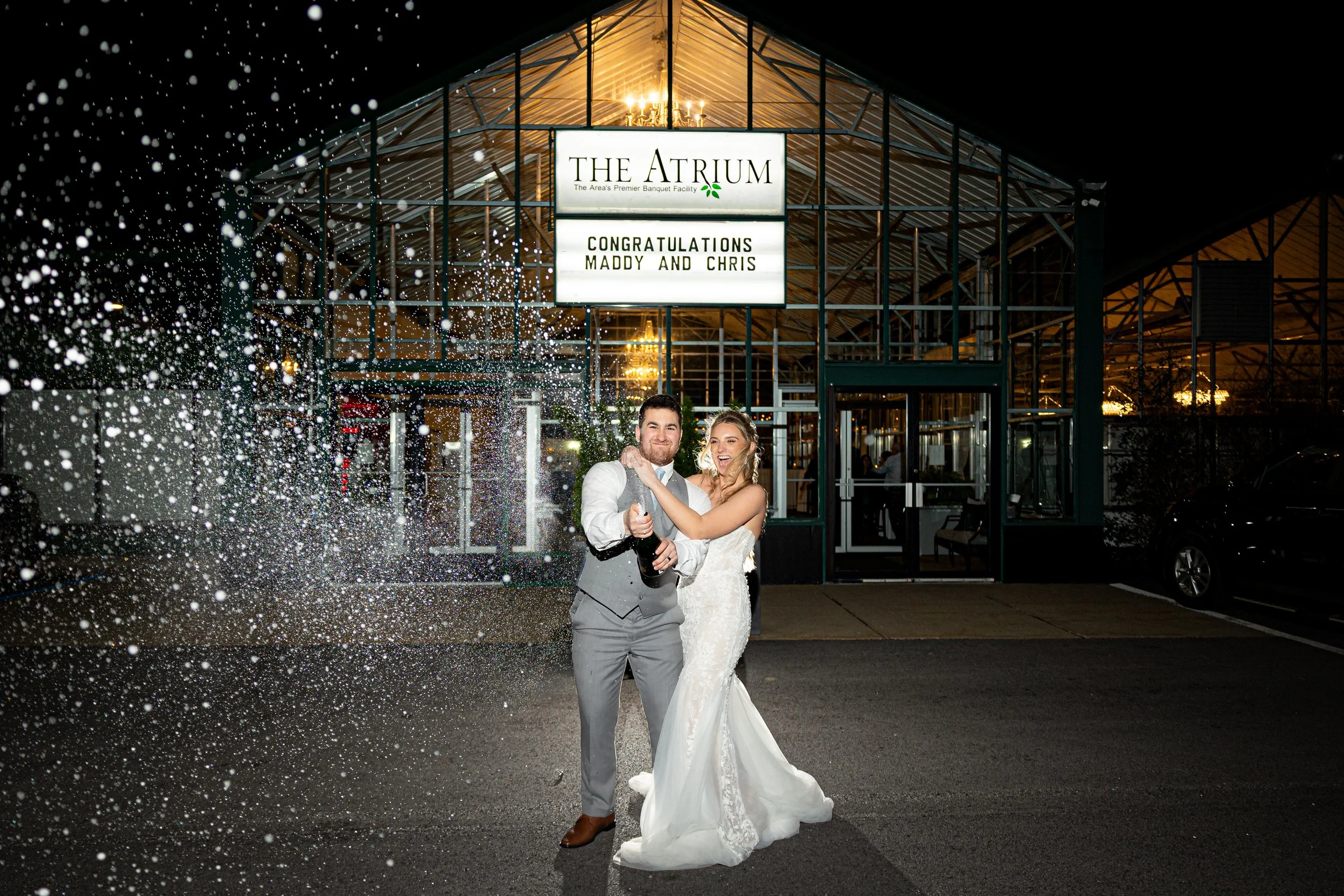 A newlywed couple celebrating outside a venue called The Atrium at night, with champagne, champagne spray, and a smiling bride and groom in wedding attire.