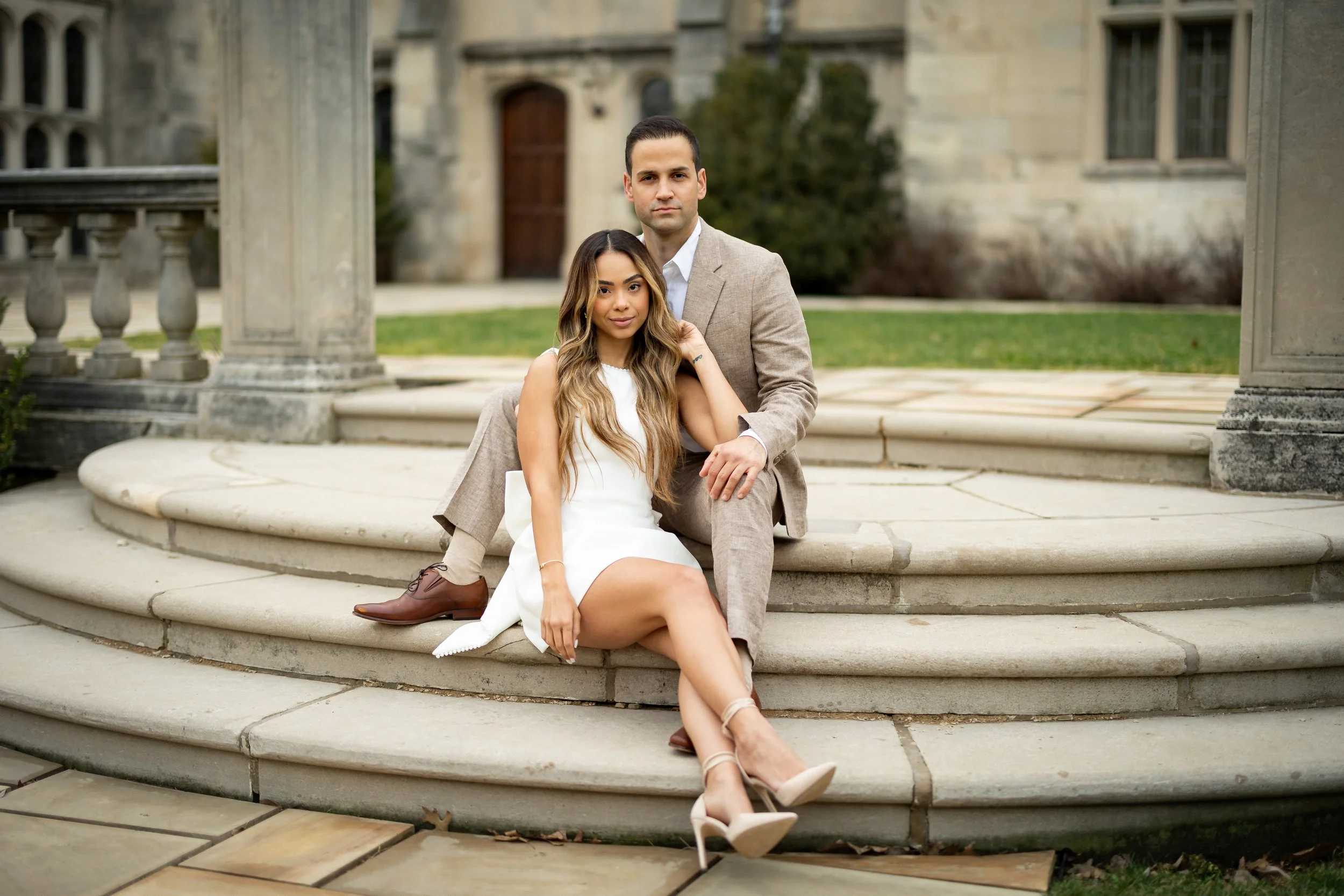 A stylishly dressed couple sitting on the steps of a stone structure outdoors, with historical building in the background.