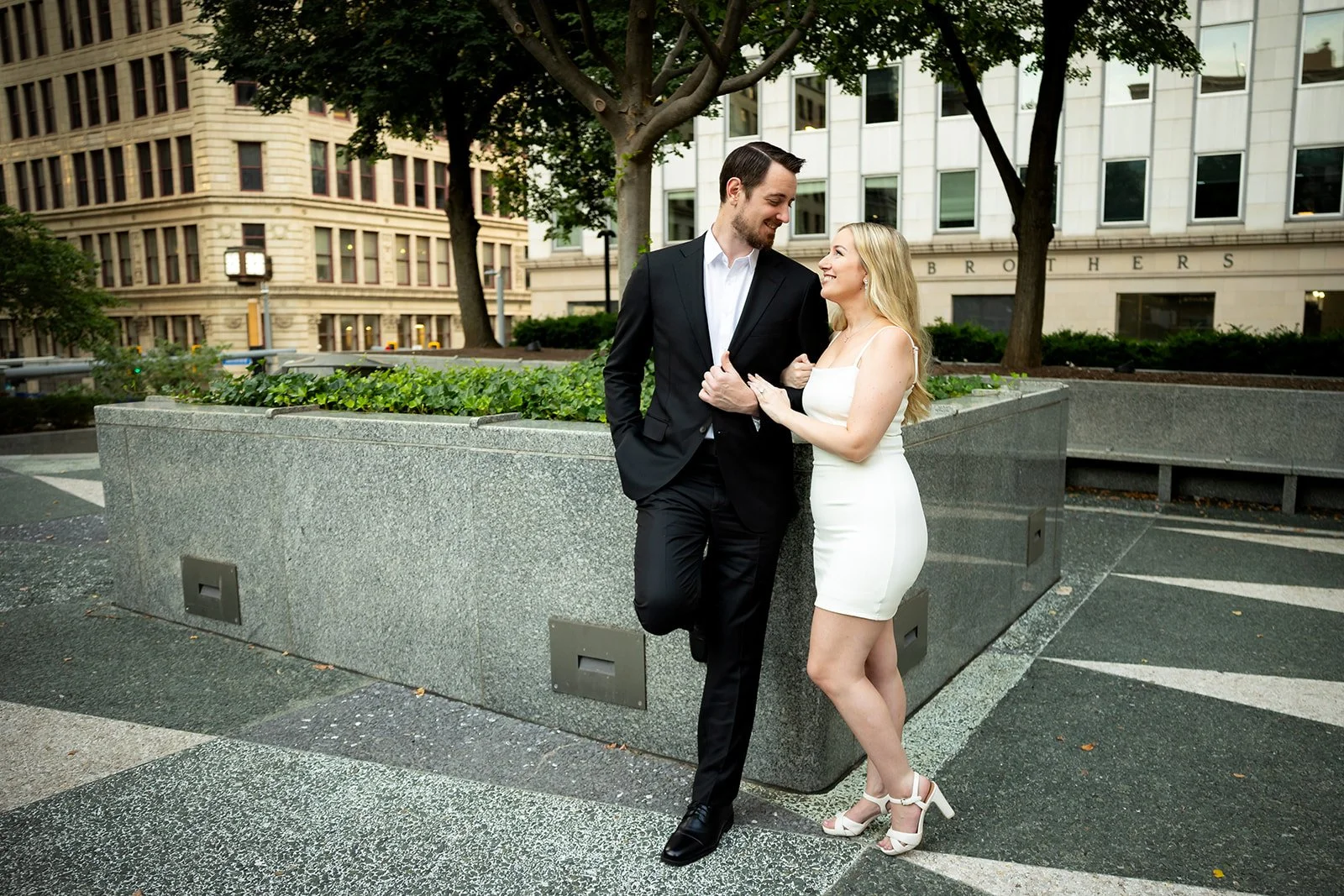 A couple, dressed in formal attire, smiling and looking at each other in an outdoor urban setting with trees and buildings in the background.