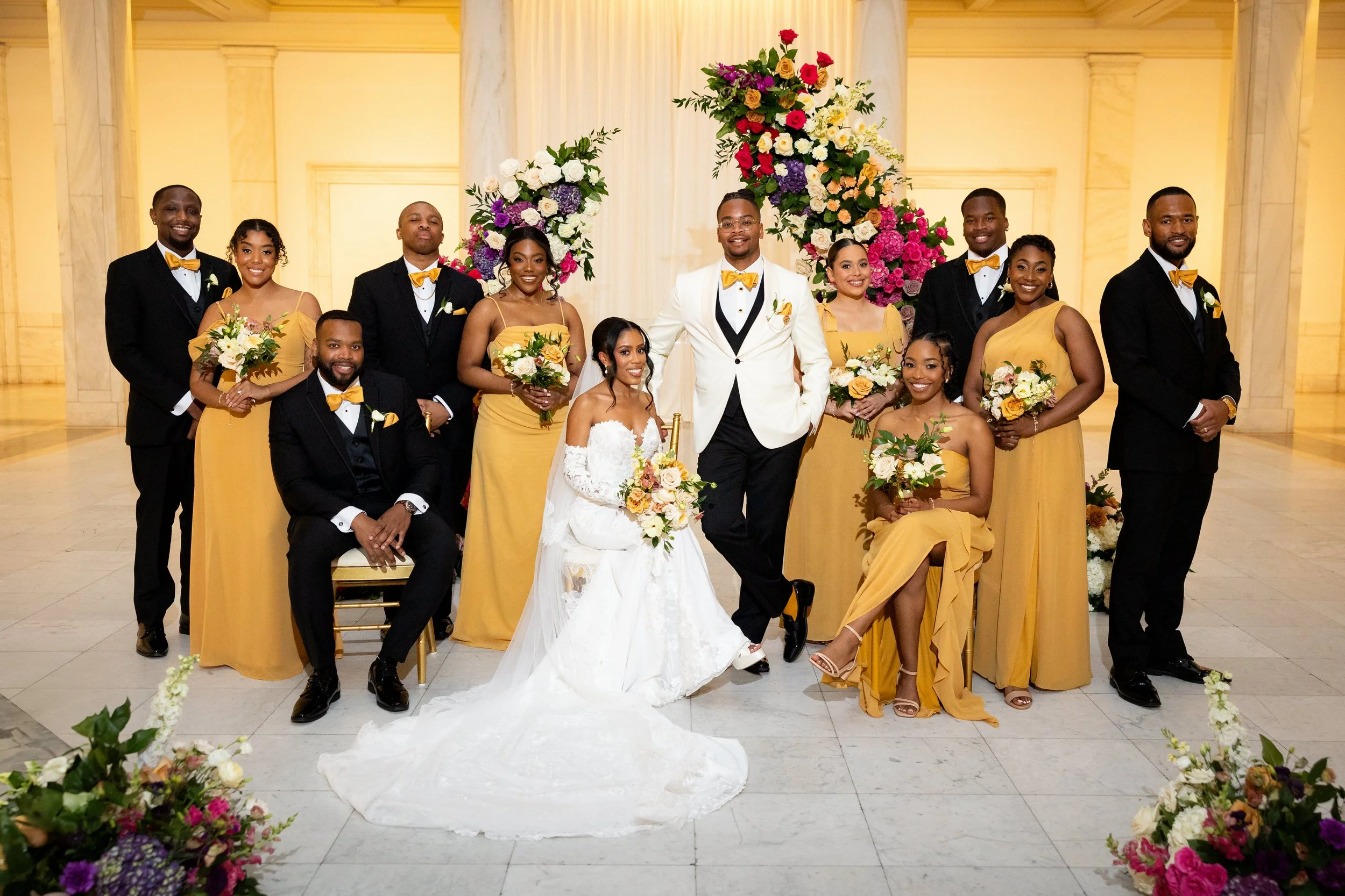 A wedding party in an elegant hall with floral arches, a bride and groom in wedding attire, and friends in matching yellow dresses and black suits, all smiling and holding bouquets.