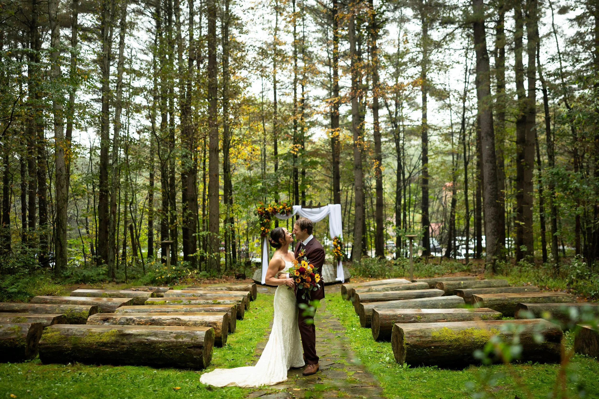A bride and groom in wedding attire share a kiss in a forested outdoor wedding ceremony area with wooden benches and a decorated arch.
