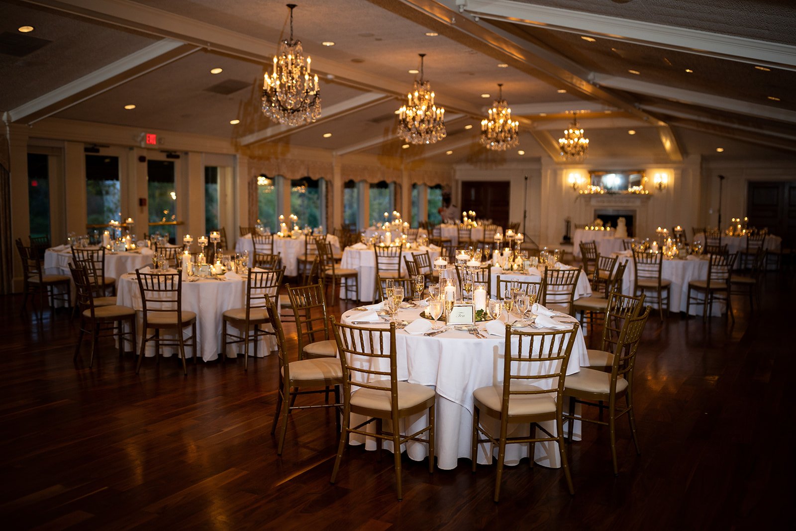 Elegant banquet hall decorated for a formal event with round tables covered in white tablecloths, surrounded by gold-colored chairs, each table set with candles, glasses, and silverware, and topped with centerpieces, illuminated by chandeliers and warm wall lighting.