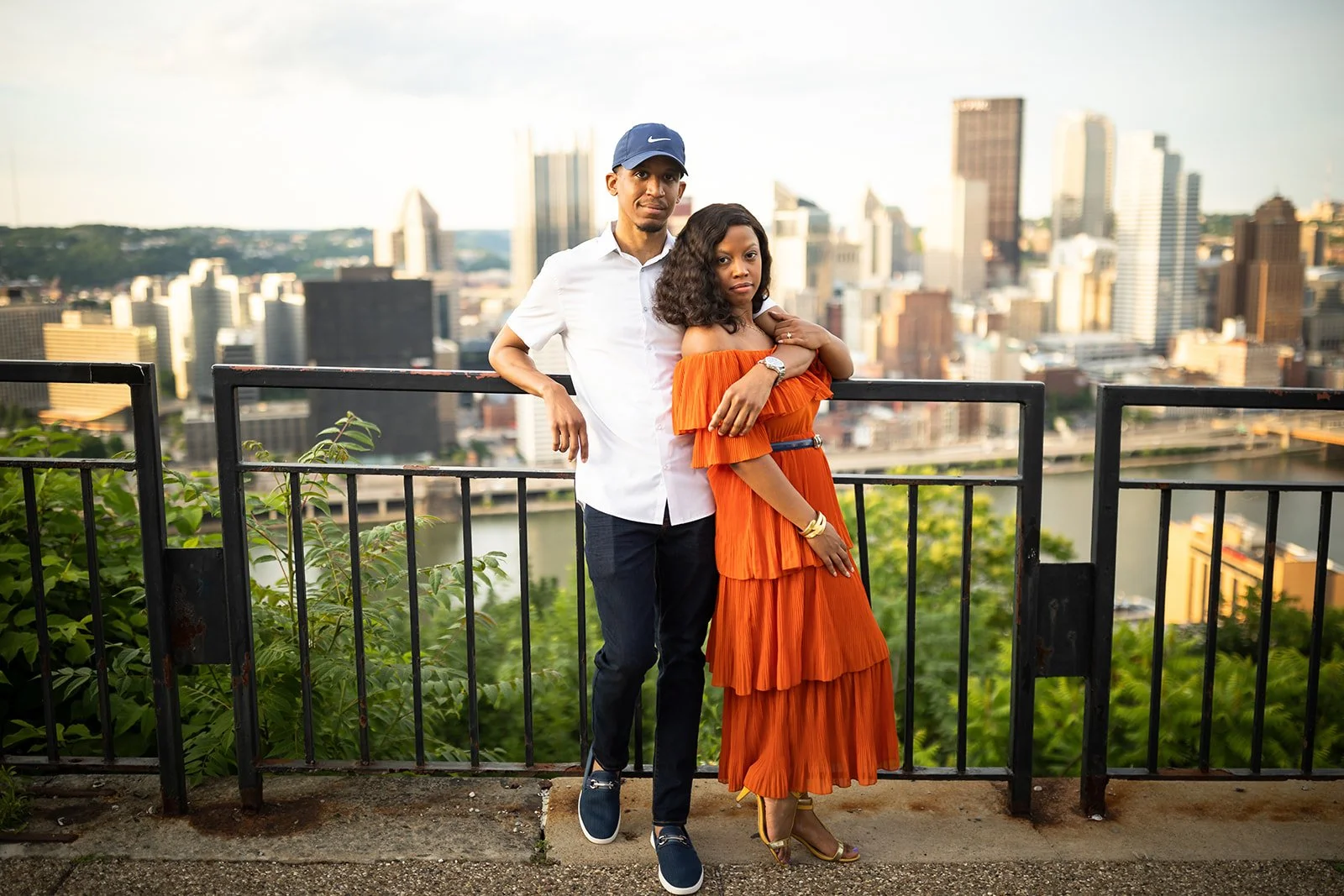 A man and woman standing on a viewing platform with a city skyline and river in the background. The man is wearing a white shirt, dark pants, a blue Nike cap, and sneakers. The woman is dressed in an orange off-the-shoulder dress with tiered ruffles and gold heels. They pose close together, with the woman holding her arm over her chest.
