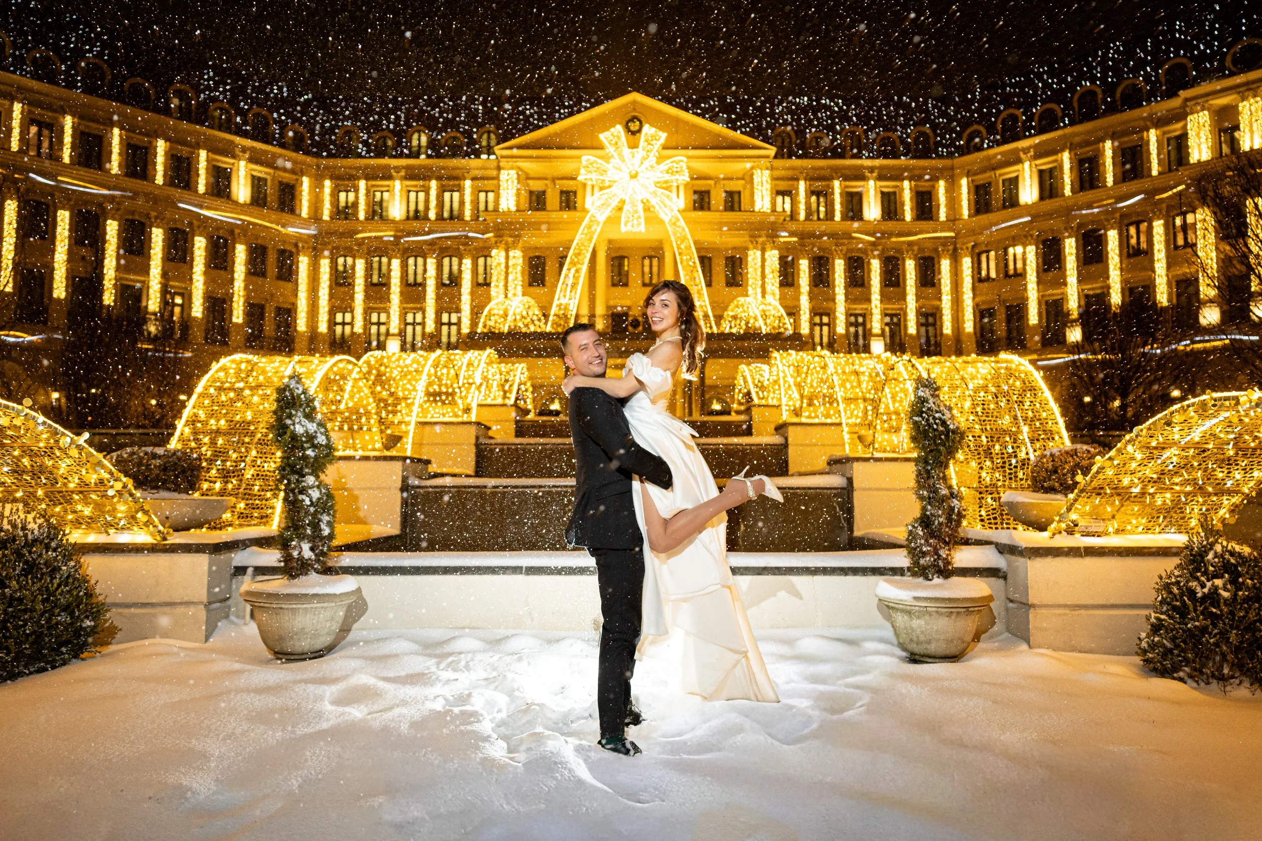 A wedding couple in wedding attire celebrates in front of a brightly decorated building with Christmas lights during a snowy night. The groom is holding the bride in his arms, and they are smiling at the camera.