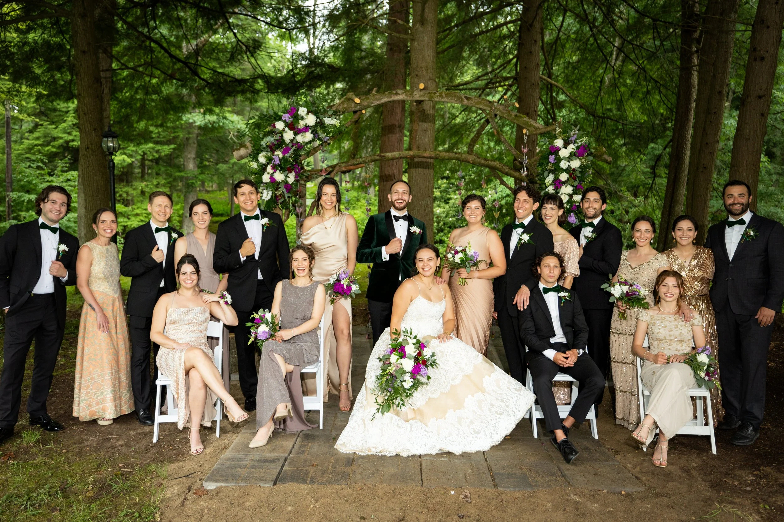 A wedding party group photo in a forest with green trees, including the bride in a white gown, the groom in a dark tuxedo, bridesmaids in pastel dresses, and groomsmen in tuxedos, positioned under a decorated floral arch.