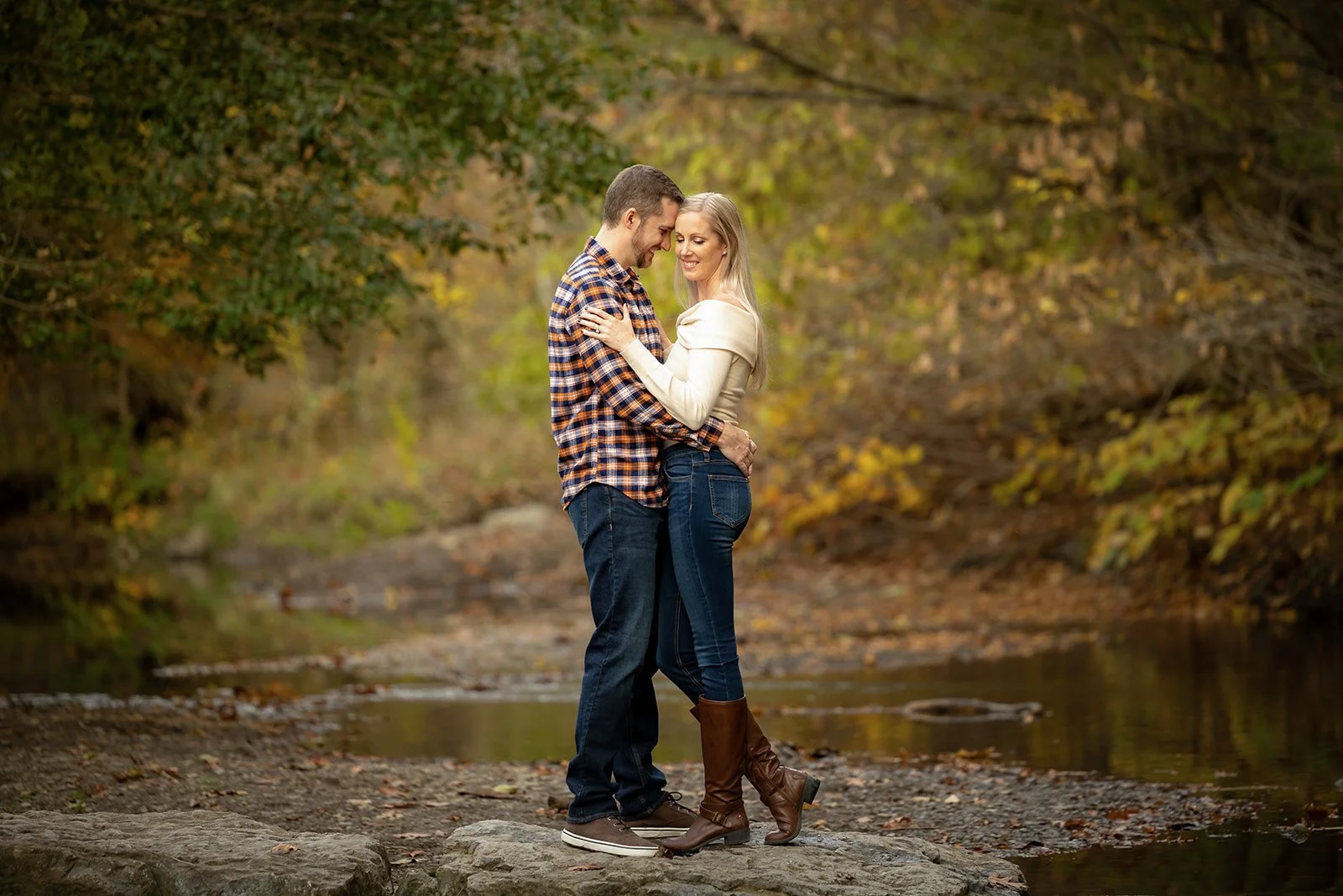 A couple embracing on a rocky waterfront with autumn trees in the background