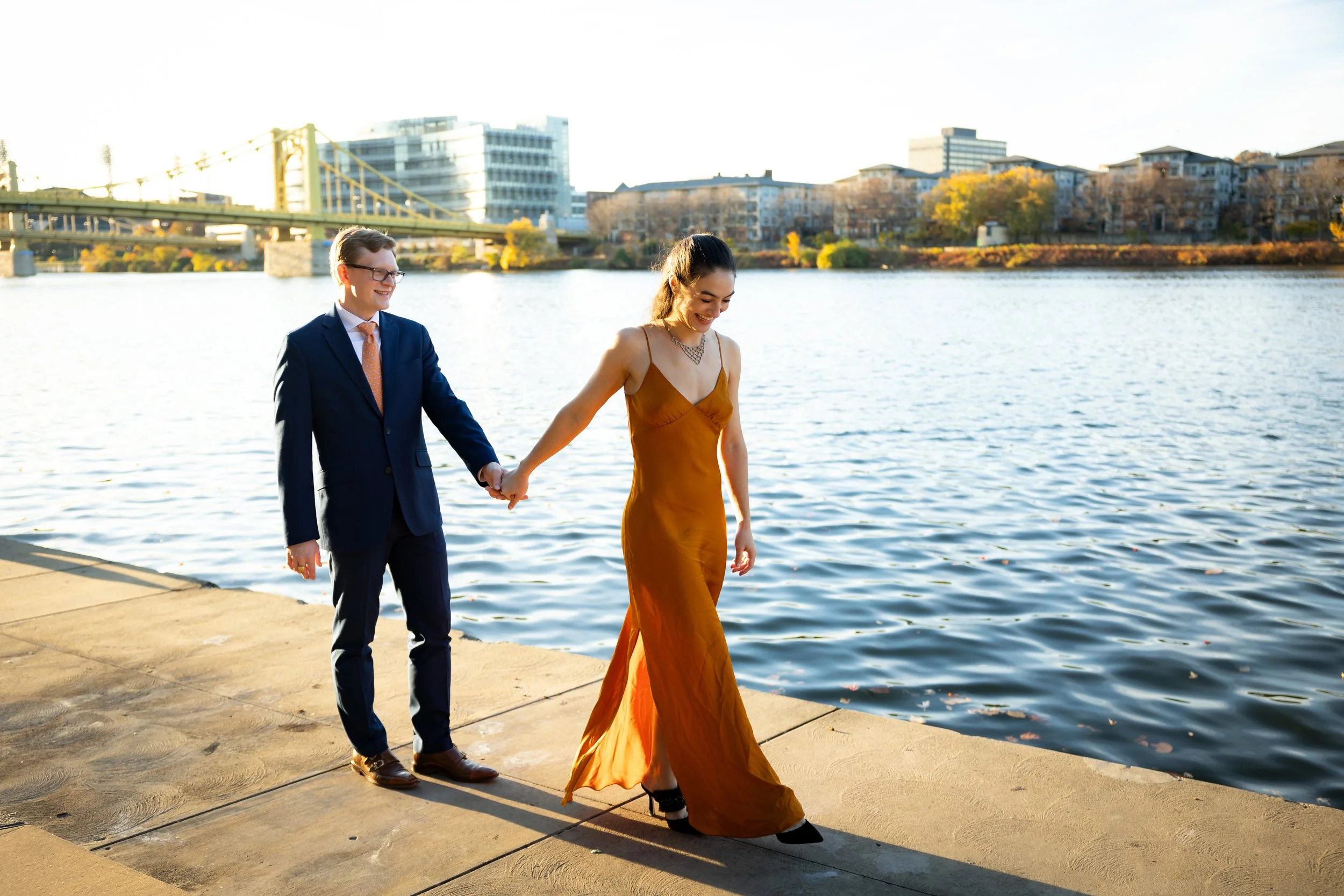 A man in a blue suit and a woman in an orange dress are holding hands and walking along a riverbank in the late afternoon. The man is smiling, and the woman is looking down and smiling. There are buildings and a bridge across the river in the background.