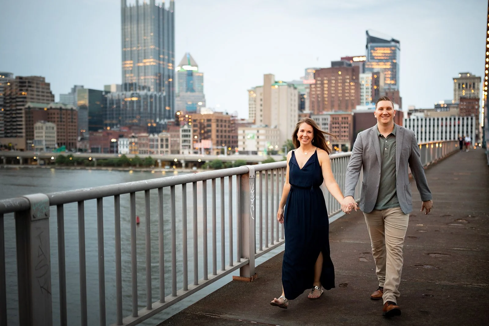 A smiling couple holding hands and walking along a city waterfront promenade with a skyline of tall buildings in the background. The woman is wearing a navy dress, and the man is in a gray blazer and khaki pants.