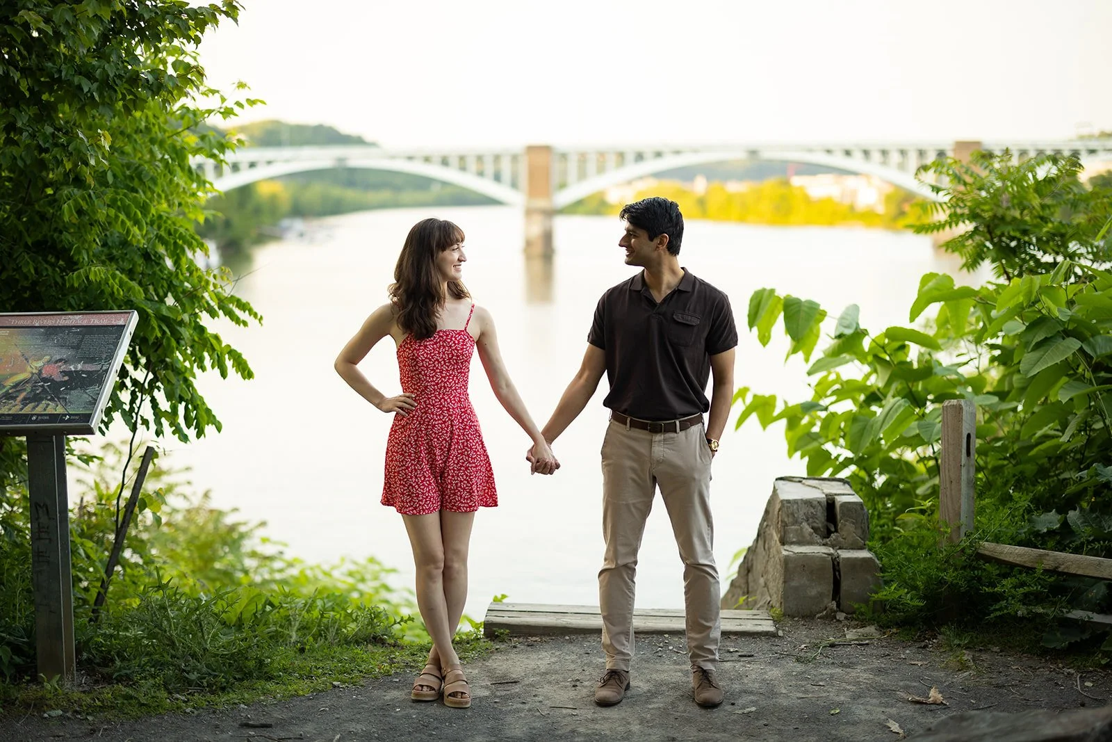 A young couple holding hands and smiling at each other near a river with a bridge in the background, surrounded by greenery.