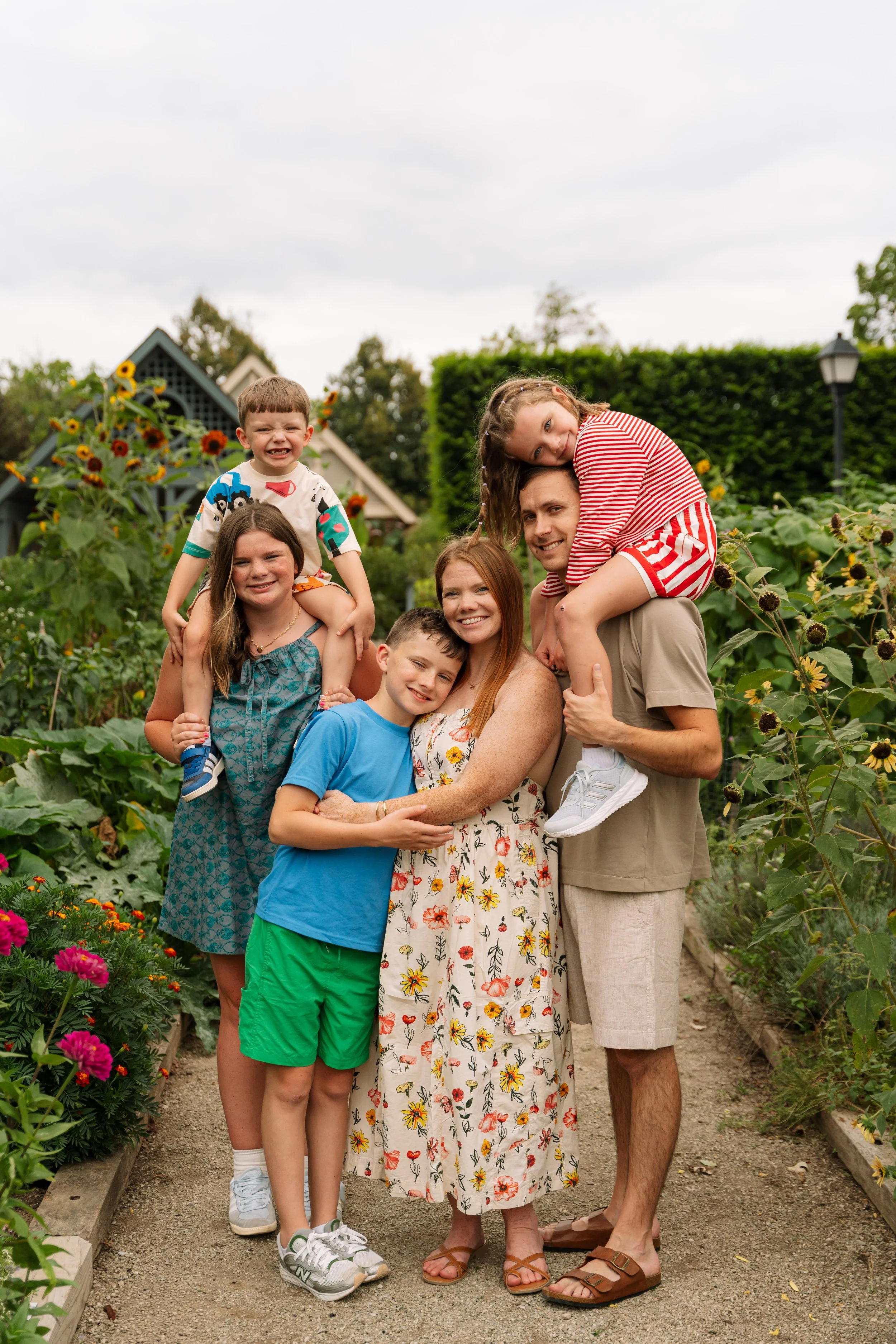 Happy family of seven poses together in a garden with sunflowers and greenery, smiling and embracing each other.