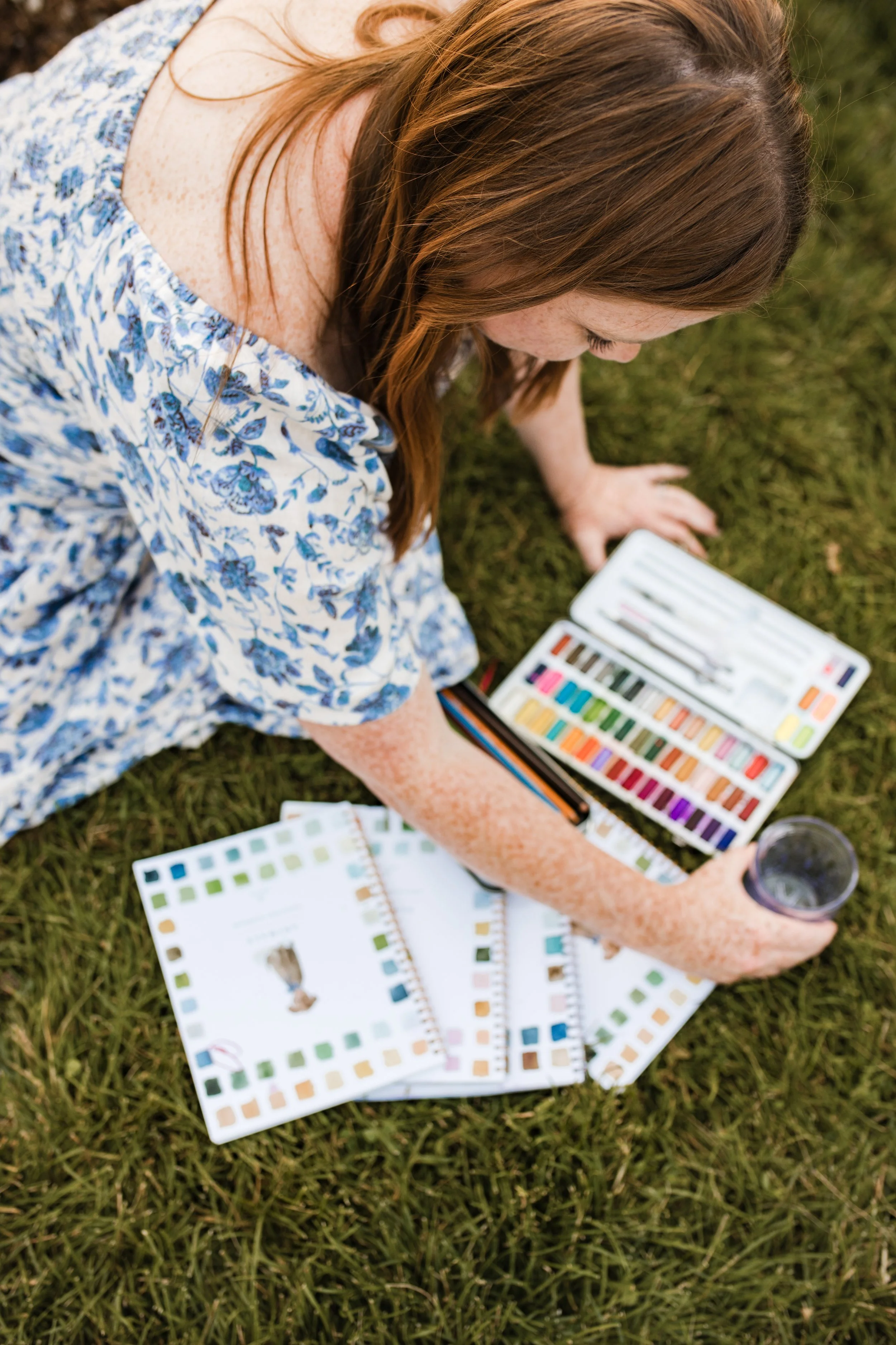A woman with red hair and freckles sitting on grass, surrounded by color swatch books, holding a glass of water, and looking at color samples.