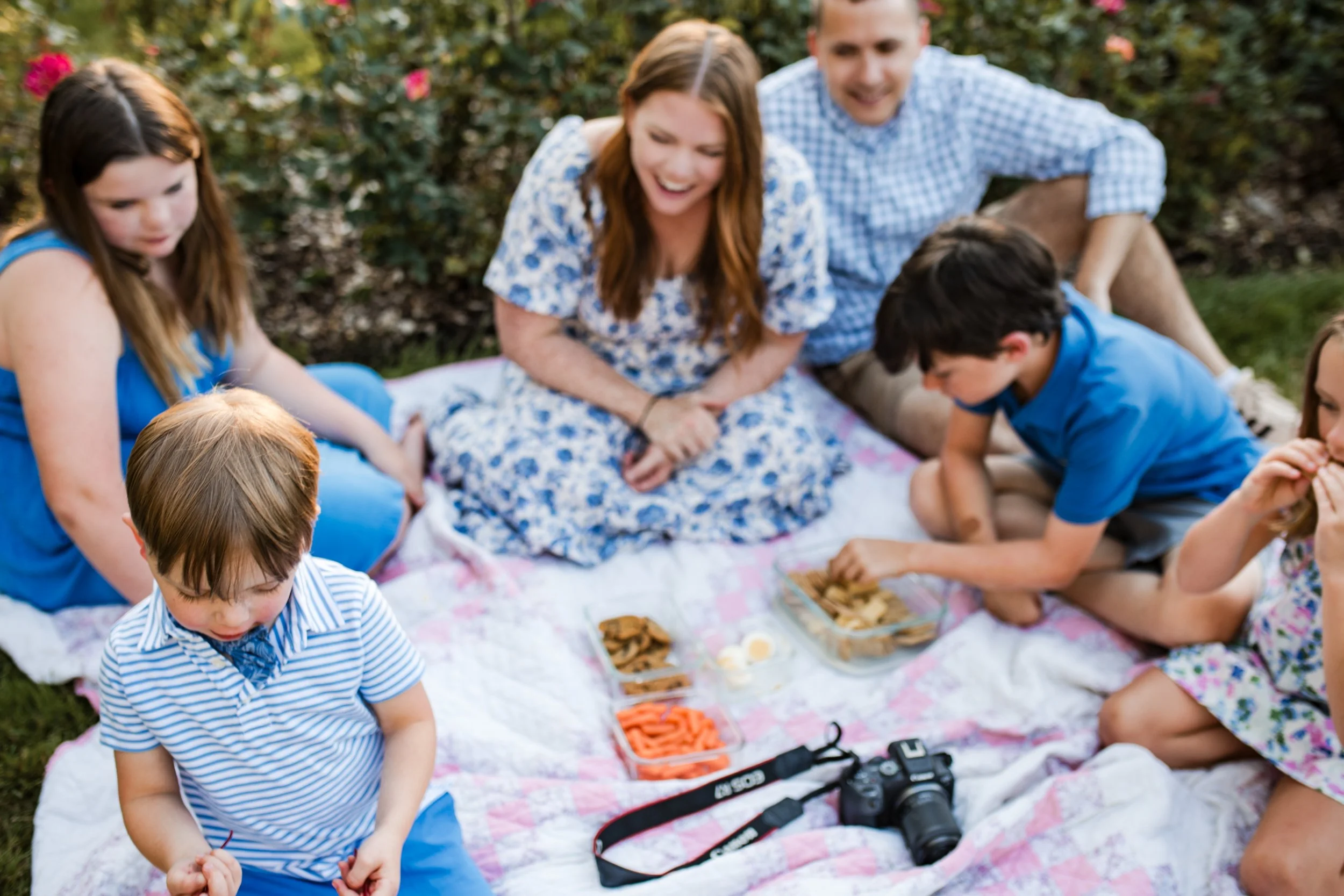 A family having a picnic outdoors on a blanket, surrounded by greenery and bushes with pink flowers, with snacks and a camera placed on the blanket.