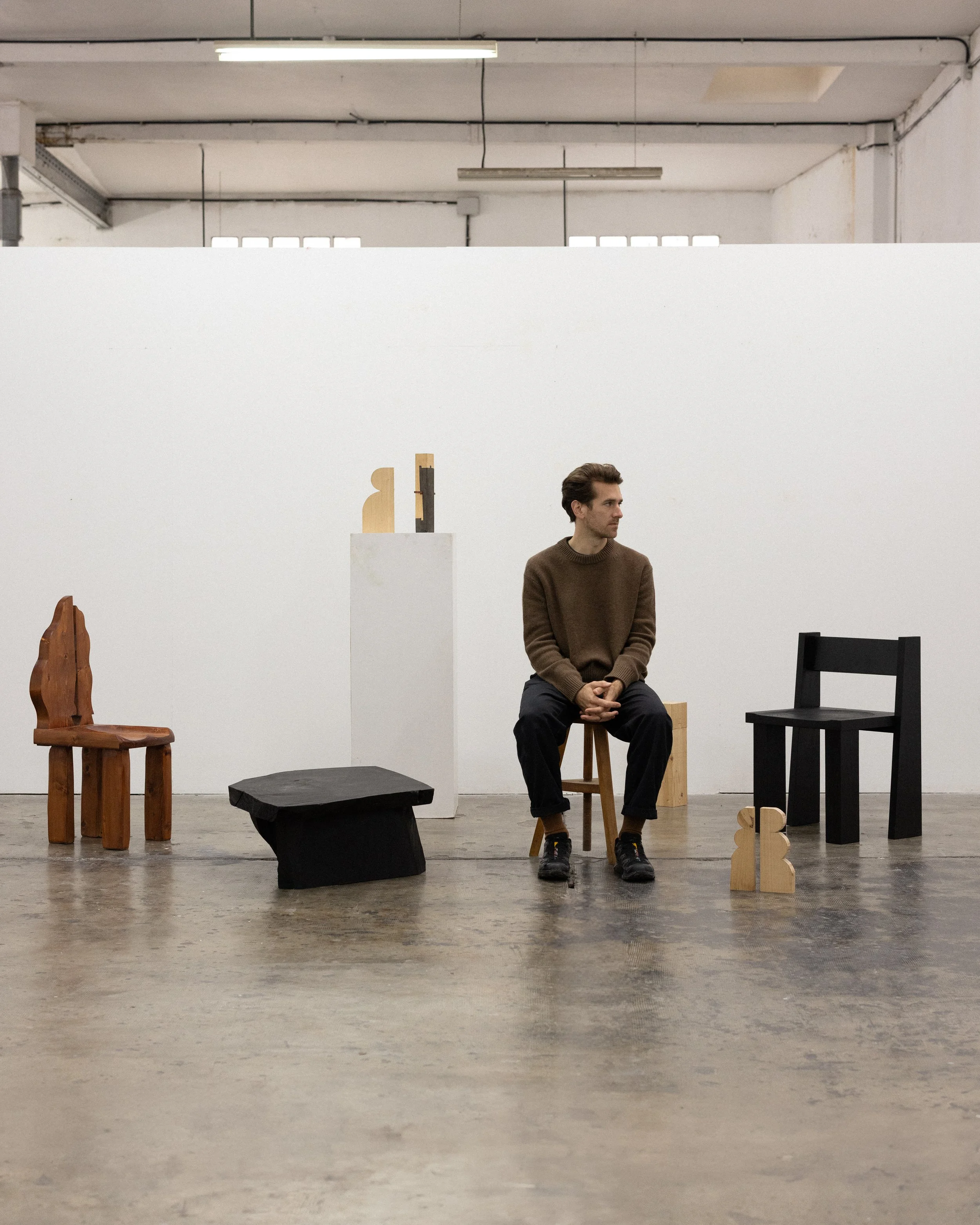 A man sits on a wooden stool in an art gallery surrounded by wooden chairs and small sculptures, with a plain white wall in the background.