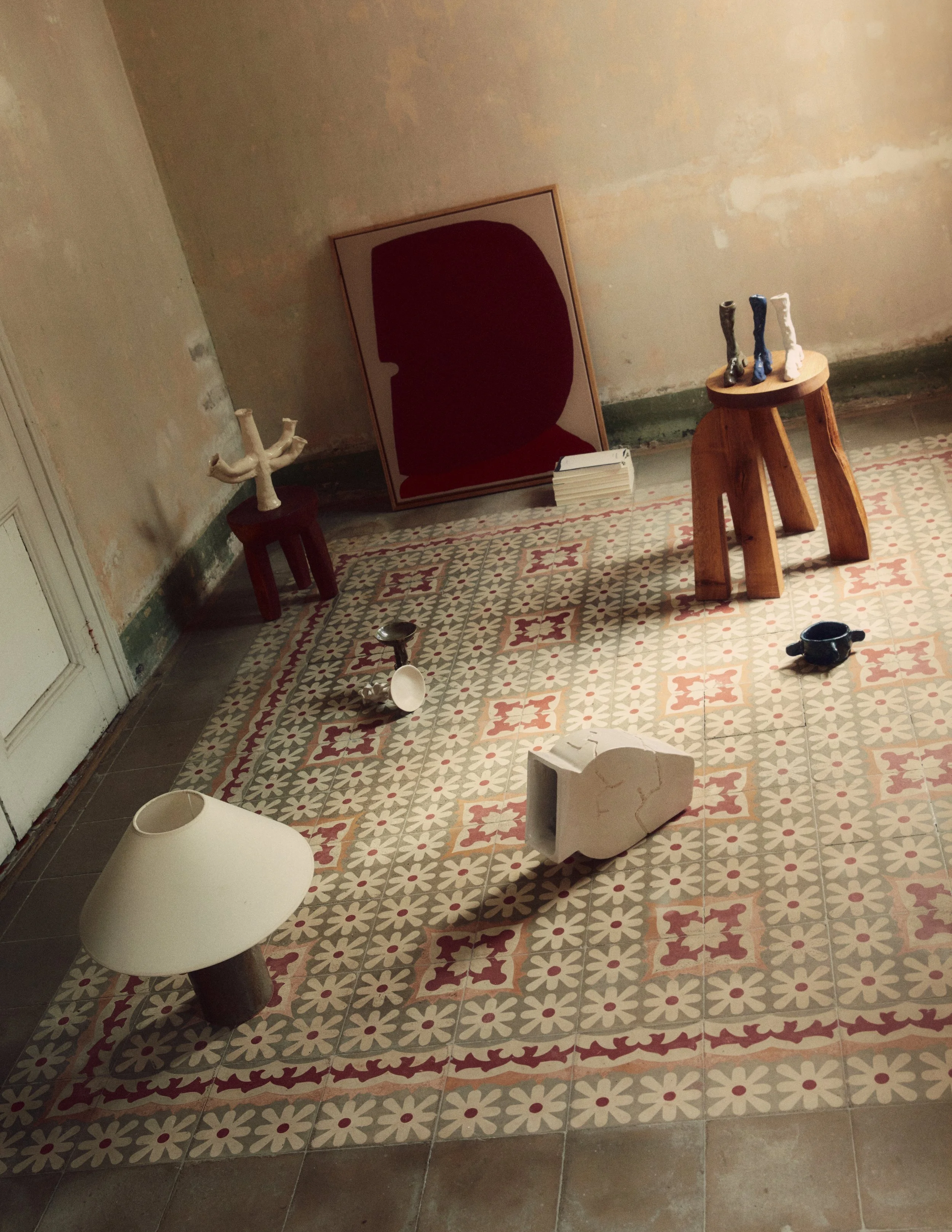Sparse room with vintage patterned tile floor, empty wall with peeling paint, and various art objects including a large red and black portrait, sculptures of feet and legs, a lamp, and small ceramic or metal bowls.