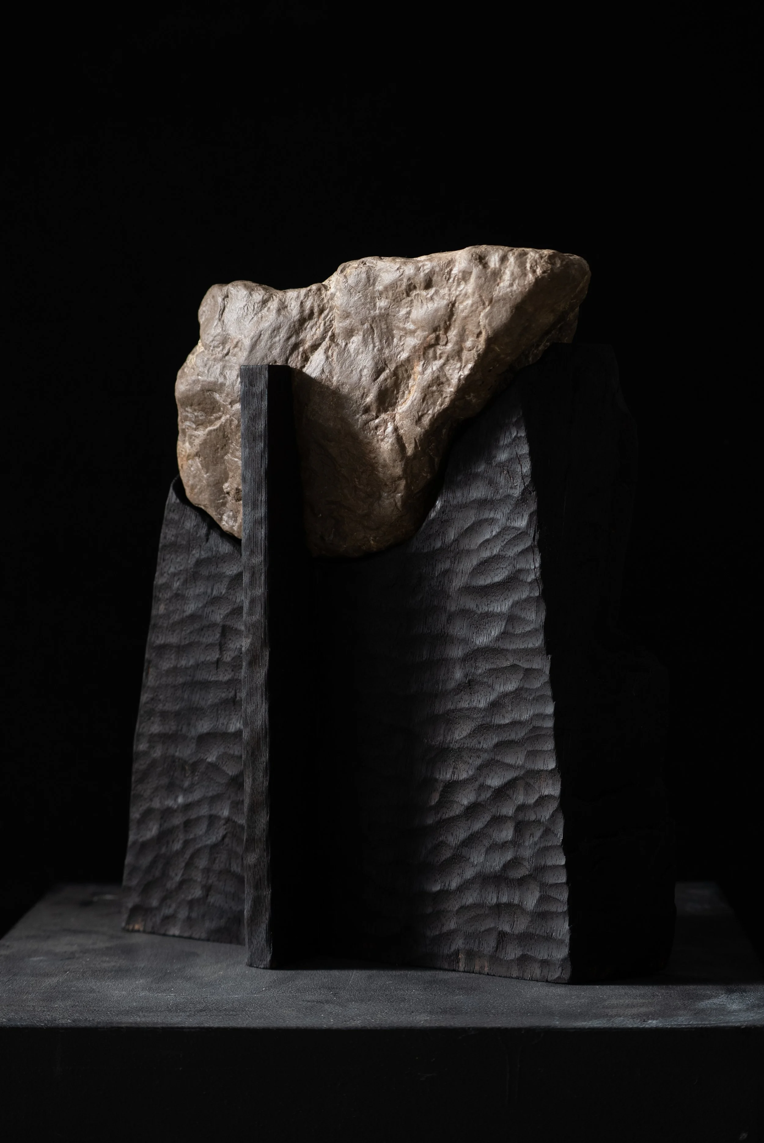 A sculpture featuring a large, jagged rock resting on a textured black wooden base with dark vertical elements against a black background.