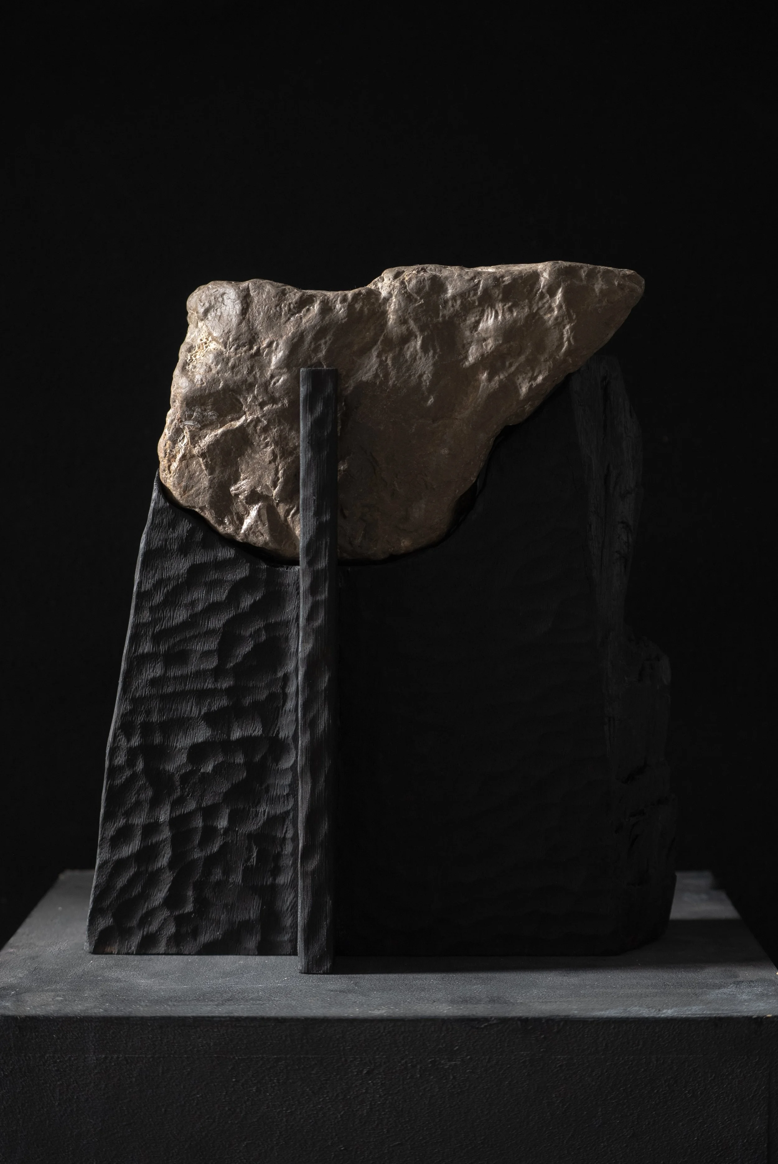 Black wooden sculpture with a rough stone on top, set against a dark background.