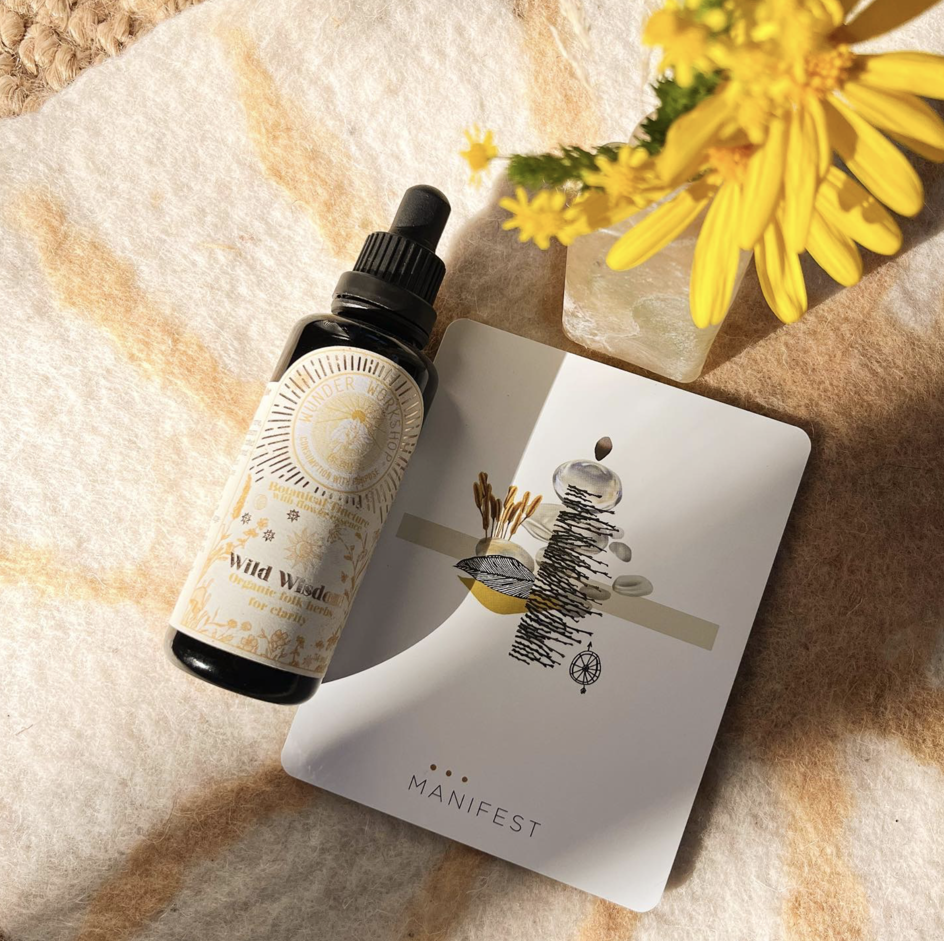 WUNDER WORKSHOP WILD WISDOM TINCTURE BOTTLE PLACED ON A TOWEL NEXT TO A HAND ILLUSTRATION AND YELLOW FLOWER