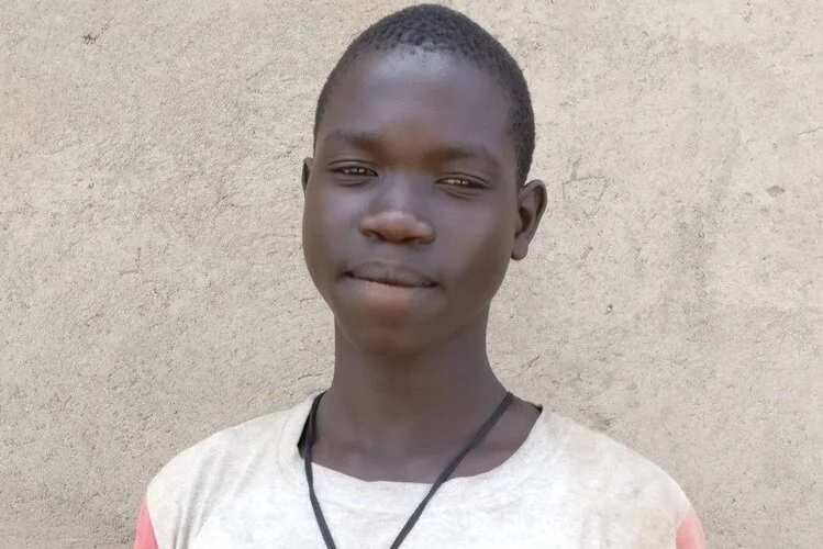 ANDREW, EL ESTUDIANTE DETERMINADO A REGRESAR A LA ESCUELA Andrew tiene 15 años y llegó a Uganda hace 6 meses después de haber huido solo de Sudán del Sur, dejando atrás a toda su familia. Andrew ahora vive solo en un asentamiento de refugiados de Palabek. Antes de que Street Child construyera la escuela primaria progresiva en Palabek, Andrew no estudiaba y no tenía nada que hacer con sus días. Sin embargo, estaba desesperado por volver a la escuela y, cuando vio la nueva escuela, se registró de inmediato. Su asignatura favorita es el inglés y en el futuro le gustaría ser cajero de un banco.