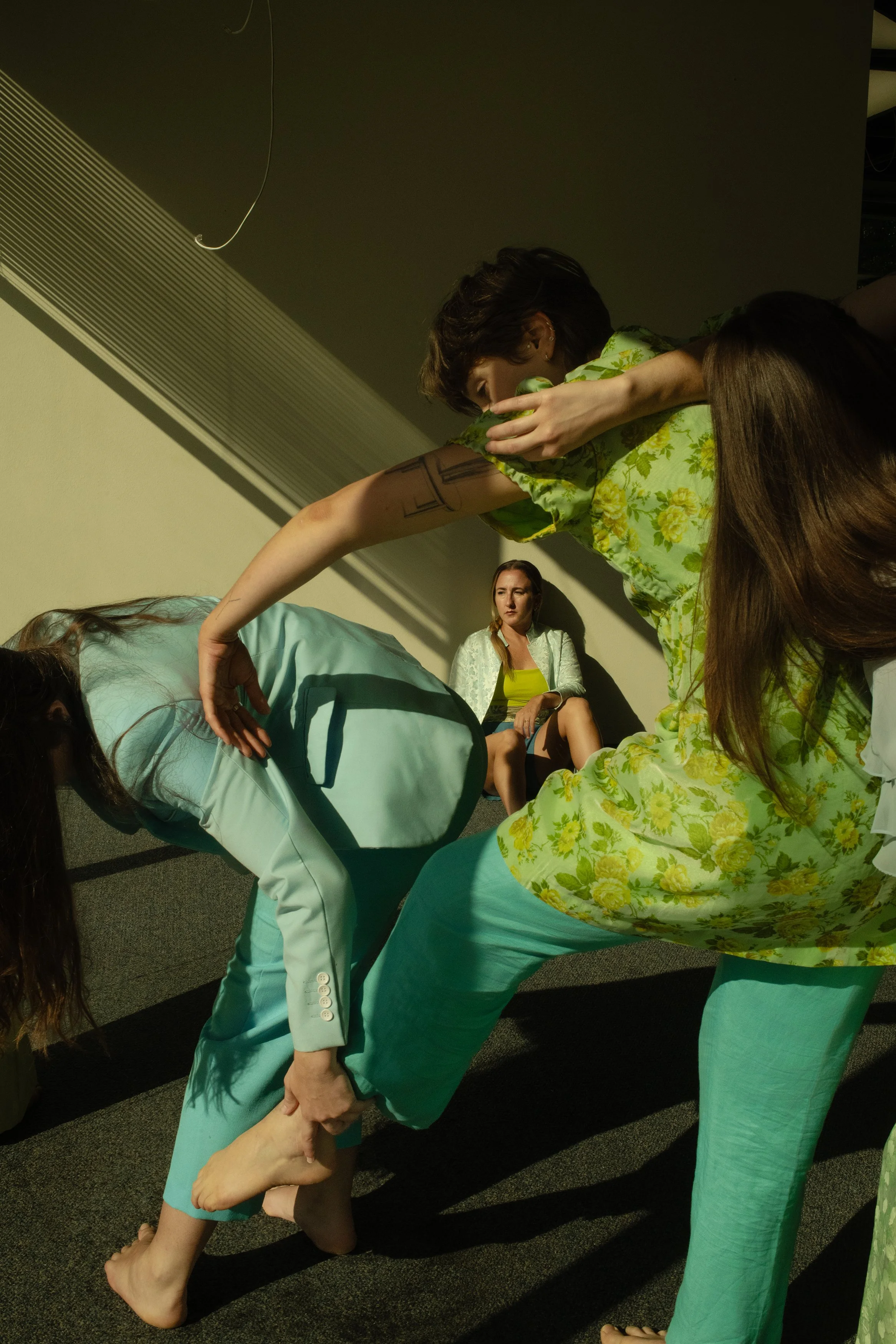 Carpeted office building. Three brunette dancers create a window with their bodies to frame a fourth dancer sitting on the floor in the background. All dancers wear shades of light blue and chartreuse green.