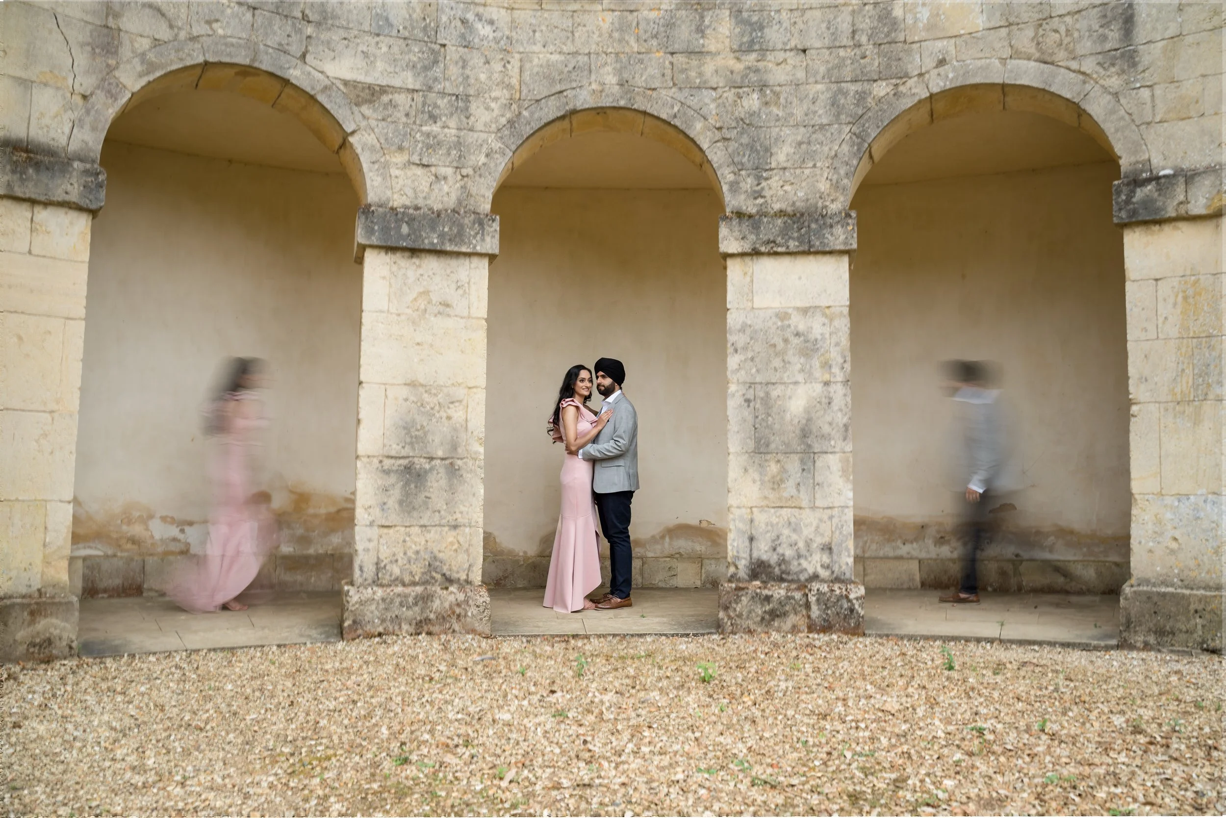 A couple stands close together, smiling at each other under historic stone arches, with two people blurred walking past on either side.