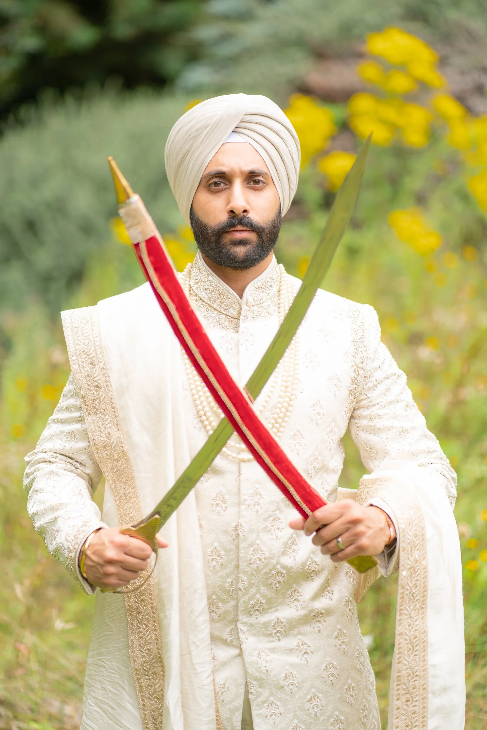 A man dressed in traditional Indian wedding attire, wearing a beige turban, holding a sword and a bow, standing outdoors in a natural setting with green foliage and yellow flowers in the background.