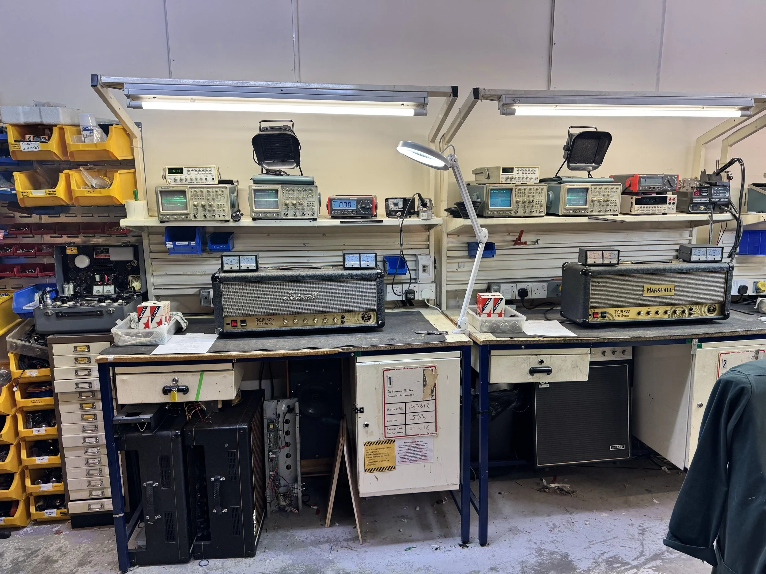 A workspace with electronic measuring equipment, electrical components, and amplifier units on a workbench in an electronics lab.
