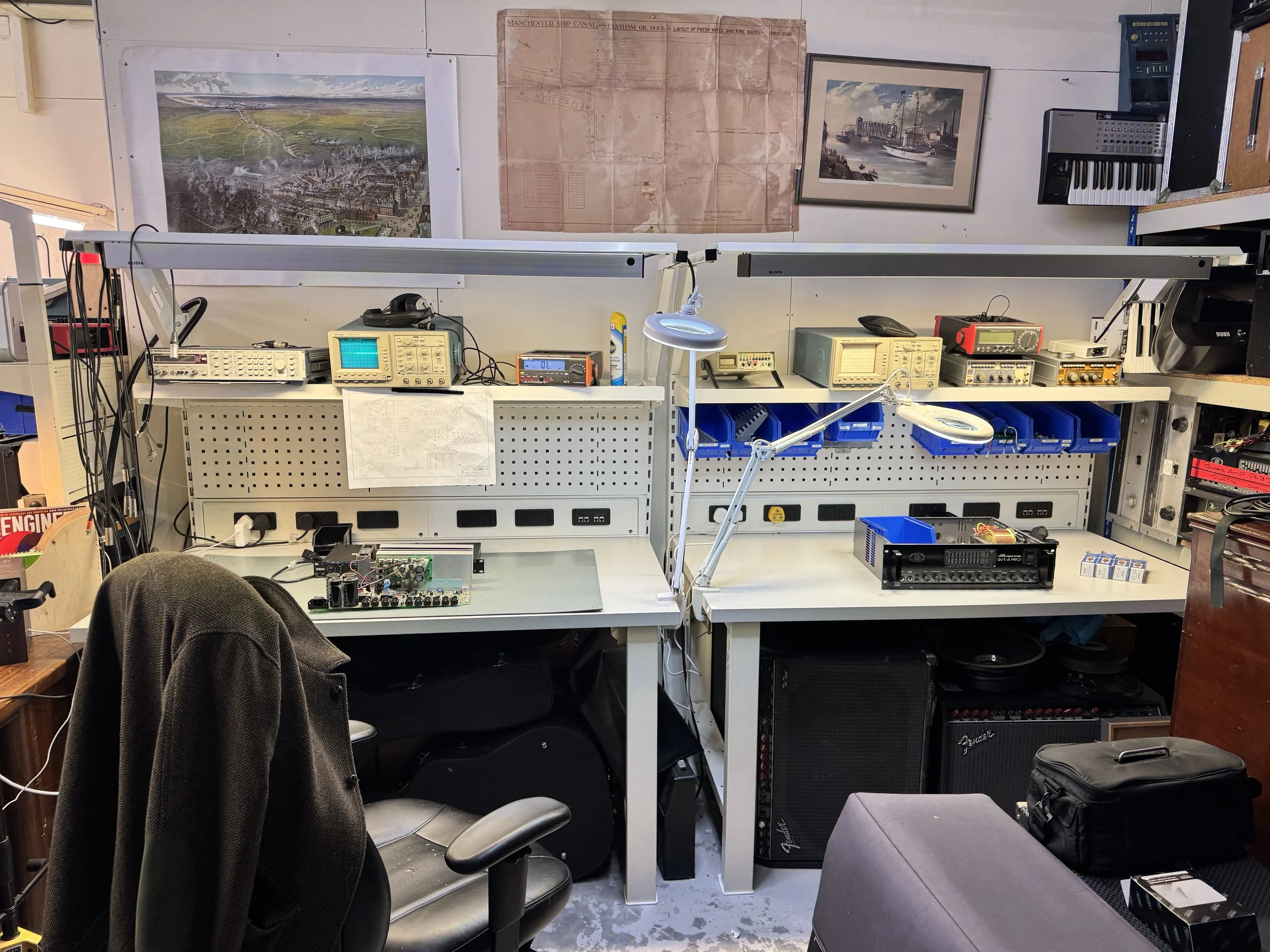 A workspace with electronic testing equipment, a computer monitor, and musical amplifier in a cluttered workshop or studio, with wall posters and framed pictures.