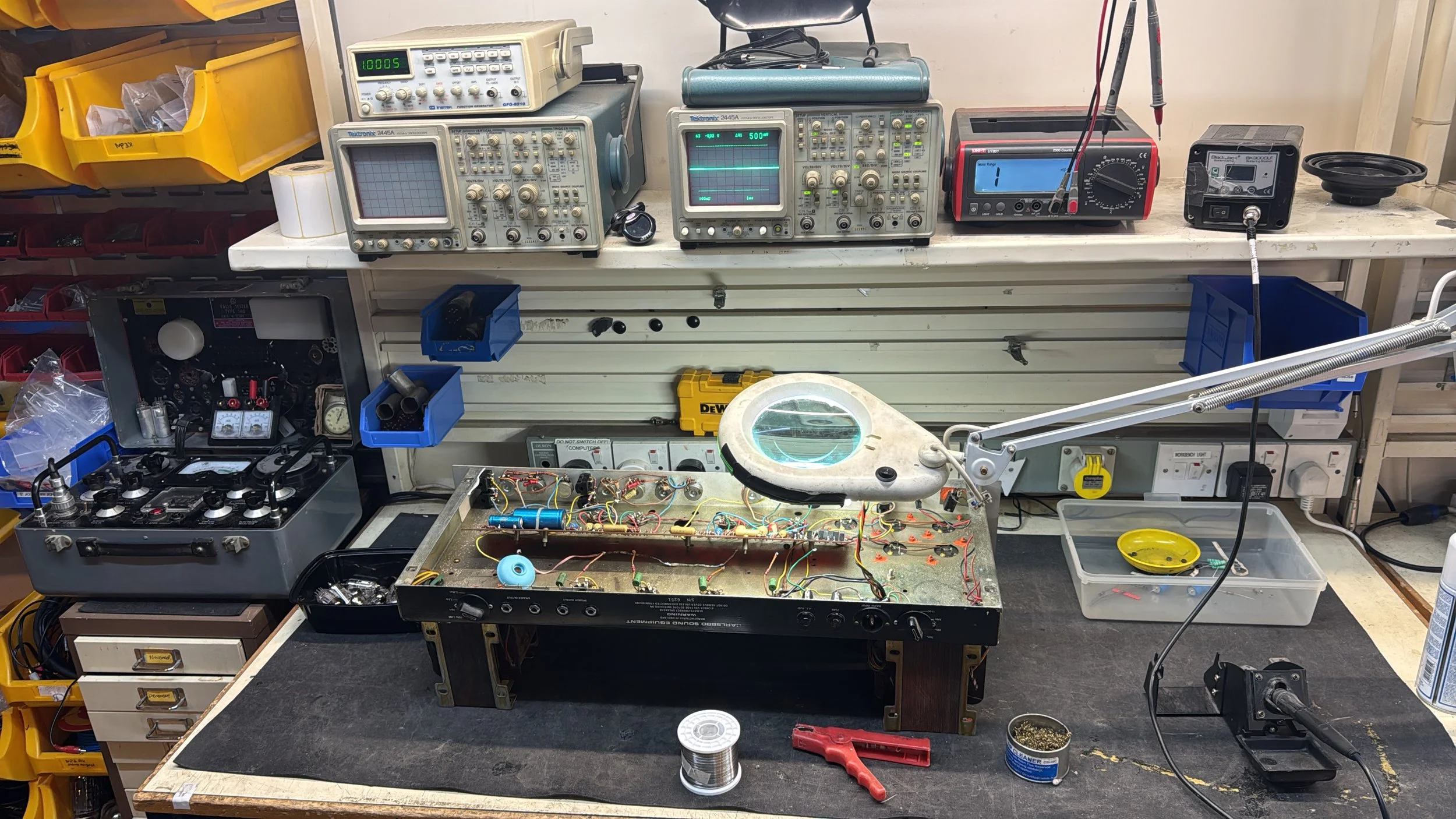 Workstation with electronic testing equipment, circuit boards, and tools for electronics repair or assembly on a workshop bench.