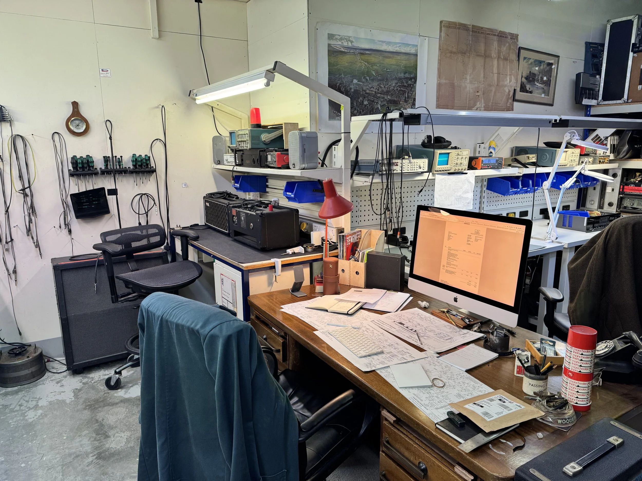 A cluttered workspace with electronic test equipment, tools, documents, and computer on a wooden desk, with wall-mounted circuit boards and framed pictures behind.