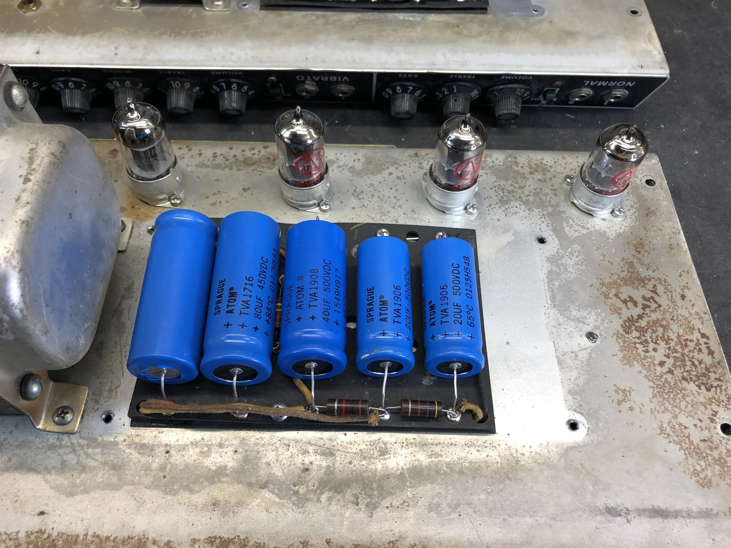 Close-up of electronic components on a circuit board, including blue cylindrical capacitors, glass vacuum tubes, resistors, and a control panel with knobs.