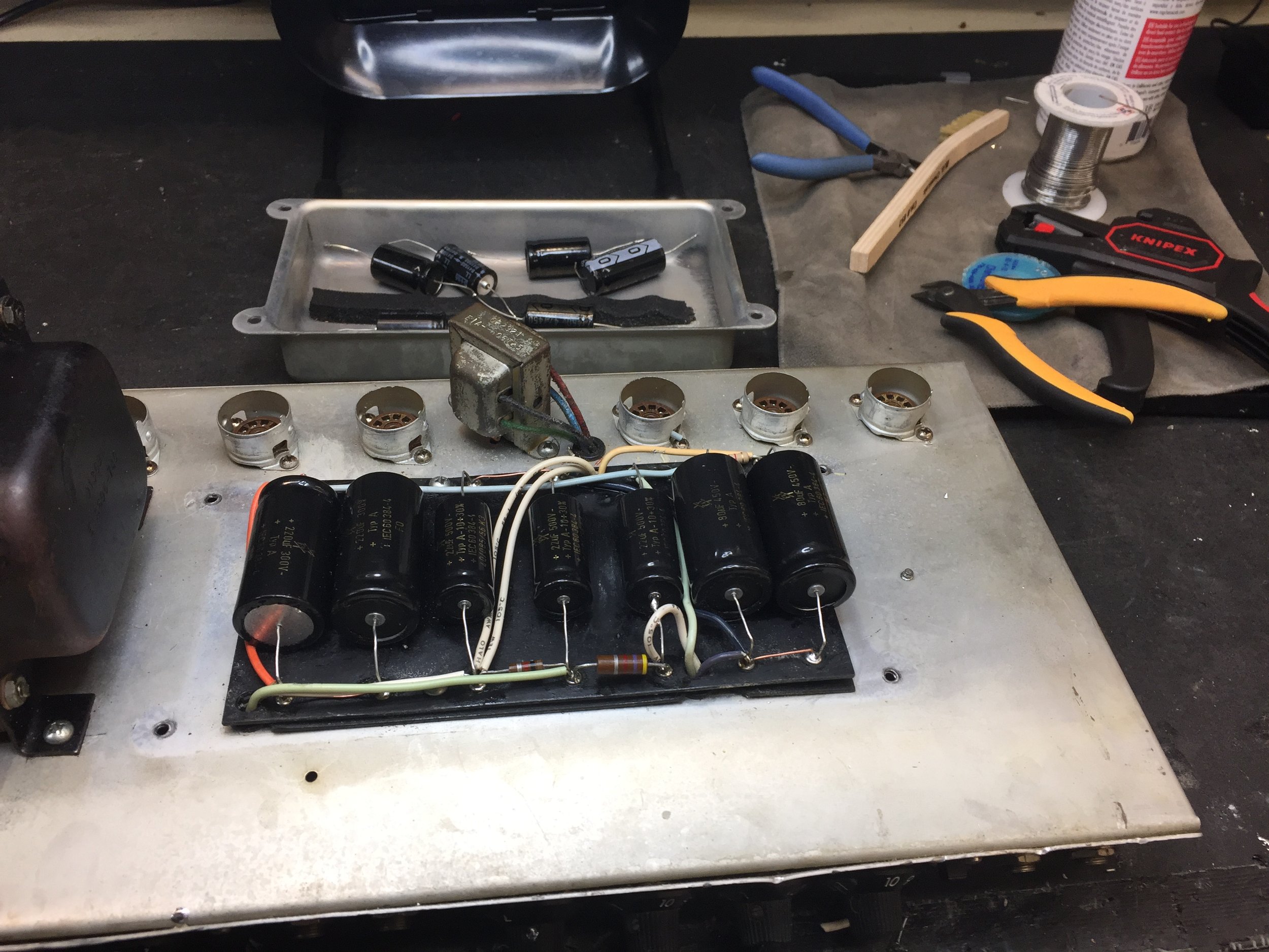 Open electronic device with capacitors, wiring, and circuit components on a workbench with tools and soldering supplies.