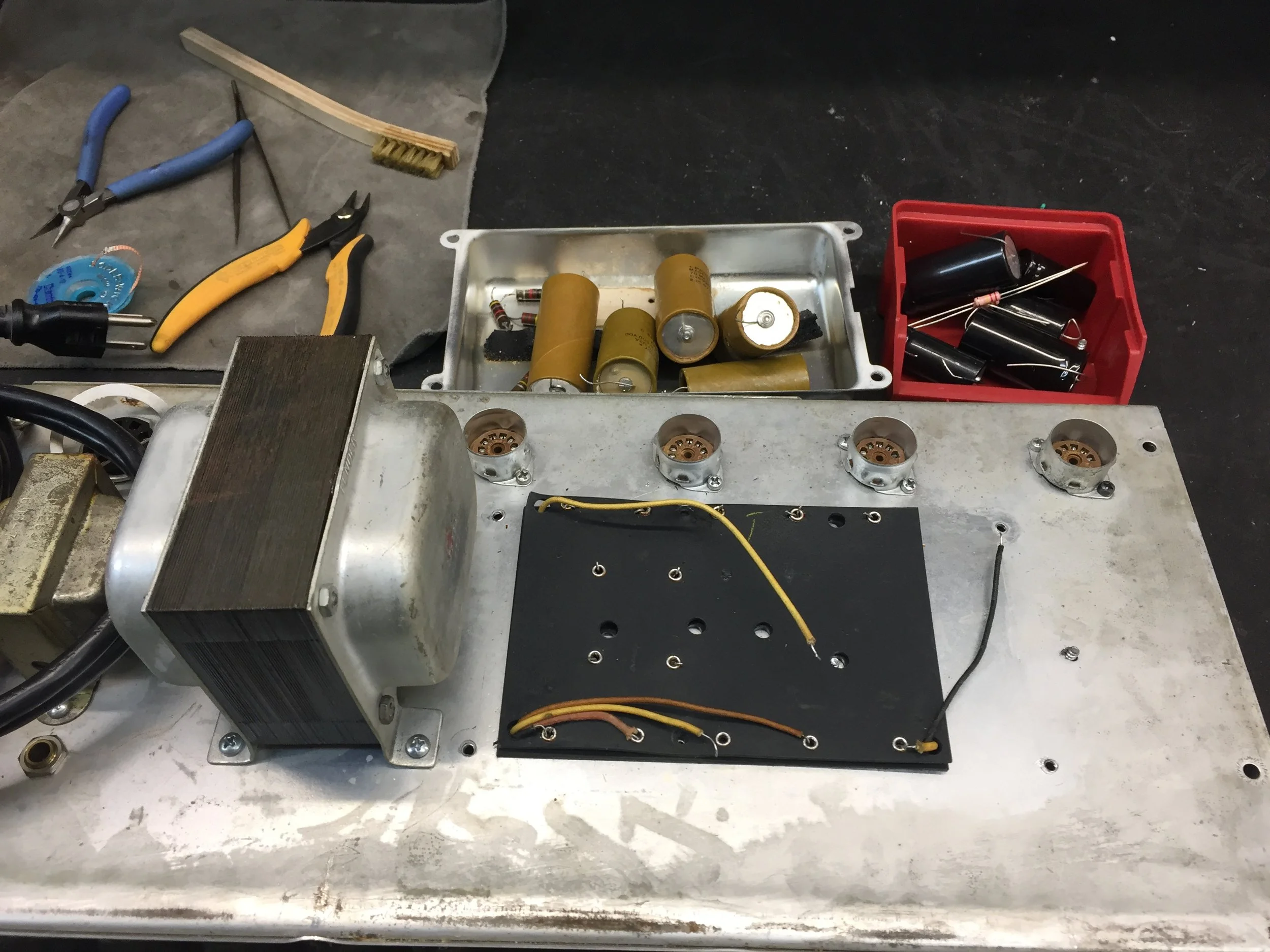 Various electronic components, wires, tools, and Capacitors on a workbench. Included are a metal enclosure with capacitors, a red plastic container with capacitors and resistors, pliers, cutting tools, a utility knife, wire, and an aluminum heatsink.