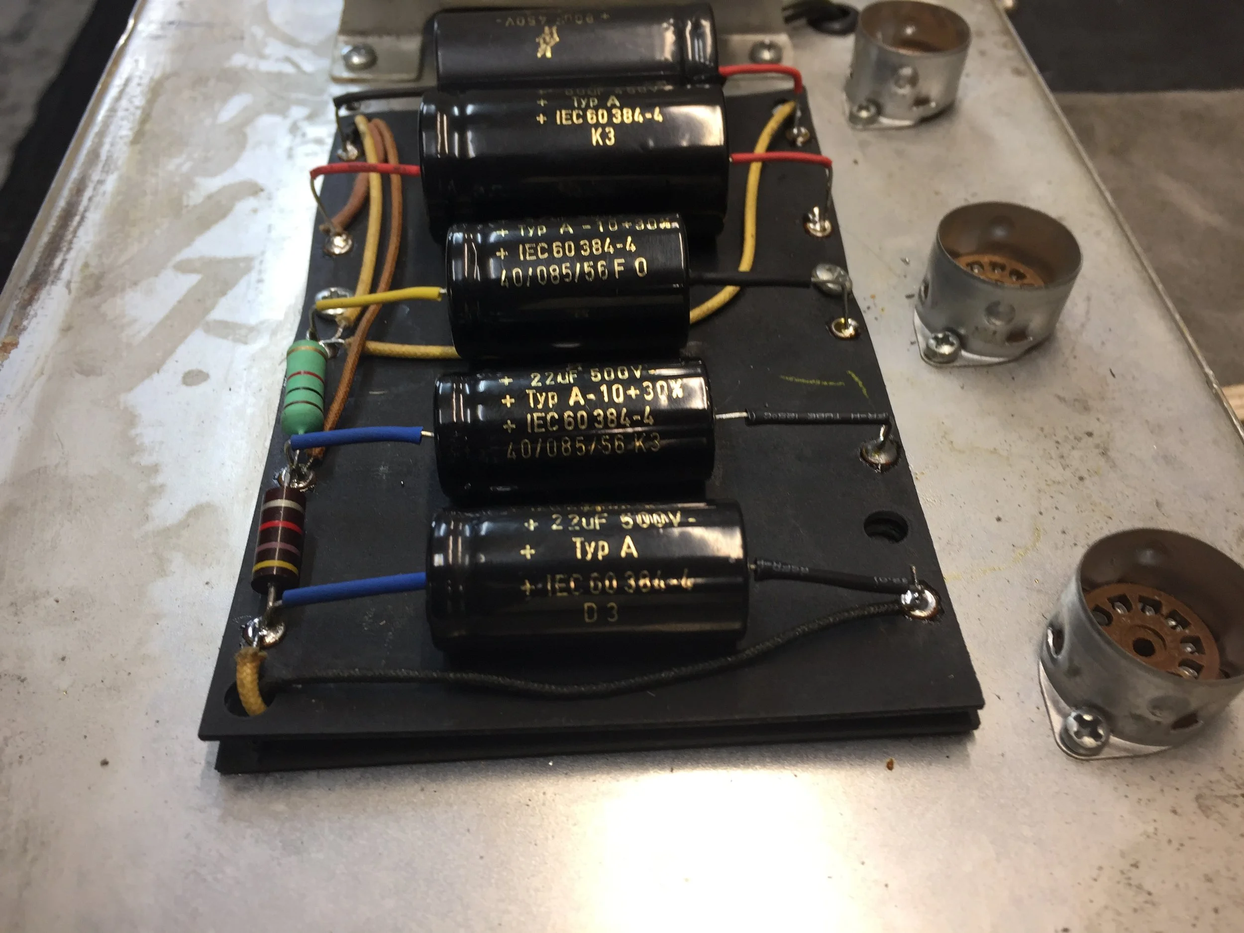 Close-up of an electronic circuit board with large black capacitors, resistors, jumper wires, and metal vacuum tubes mounted on a metal surface.