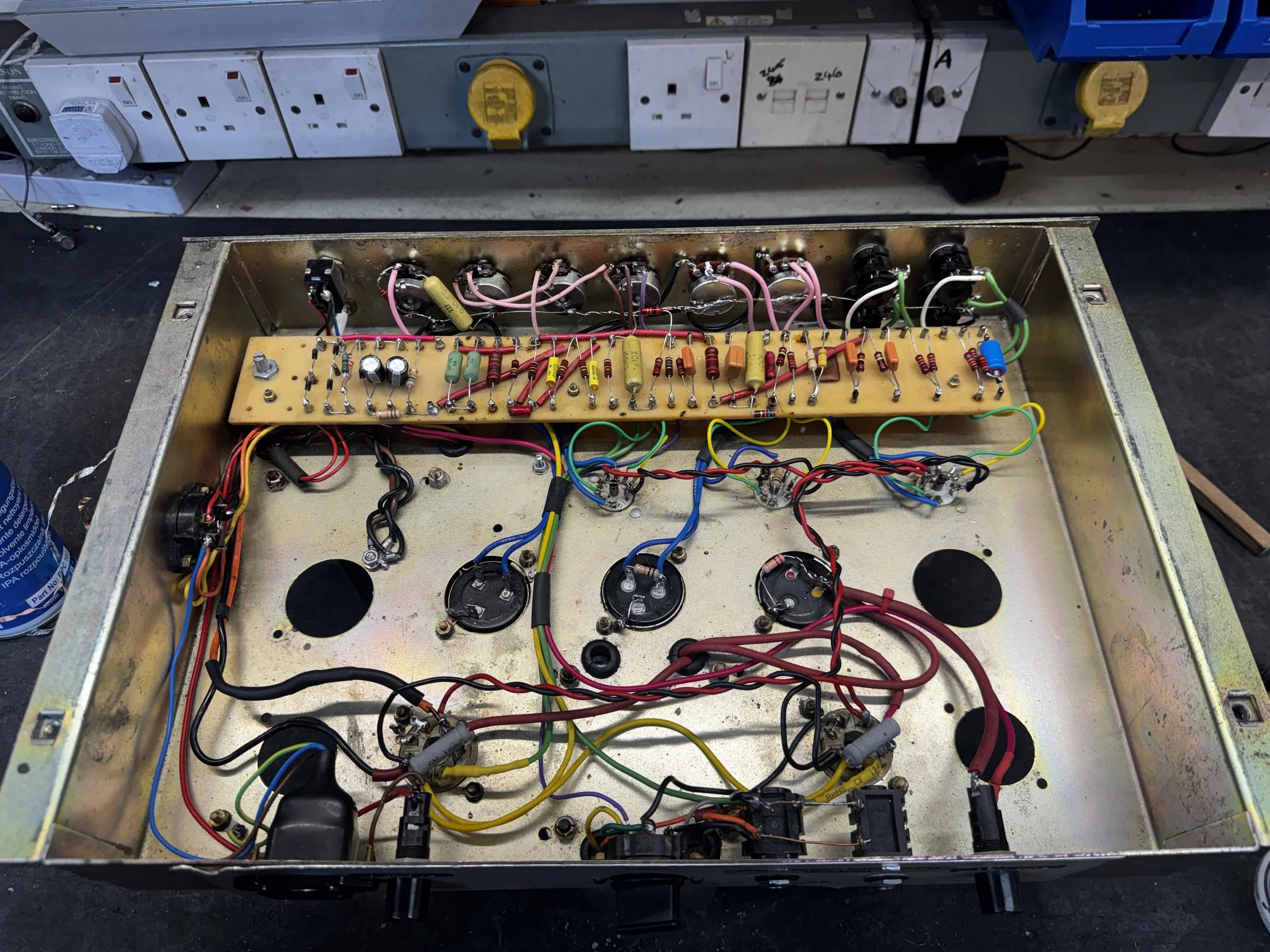 Inside view of an open electronic device, showing a circuit board with wires, resistors, and capacitors, and a row of electrical outlets and switches in the background.