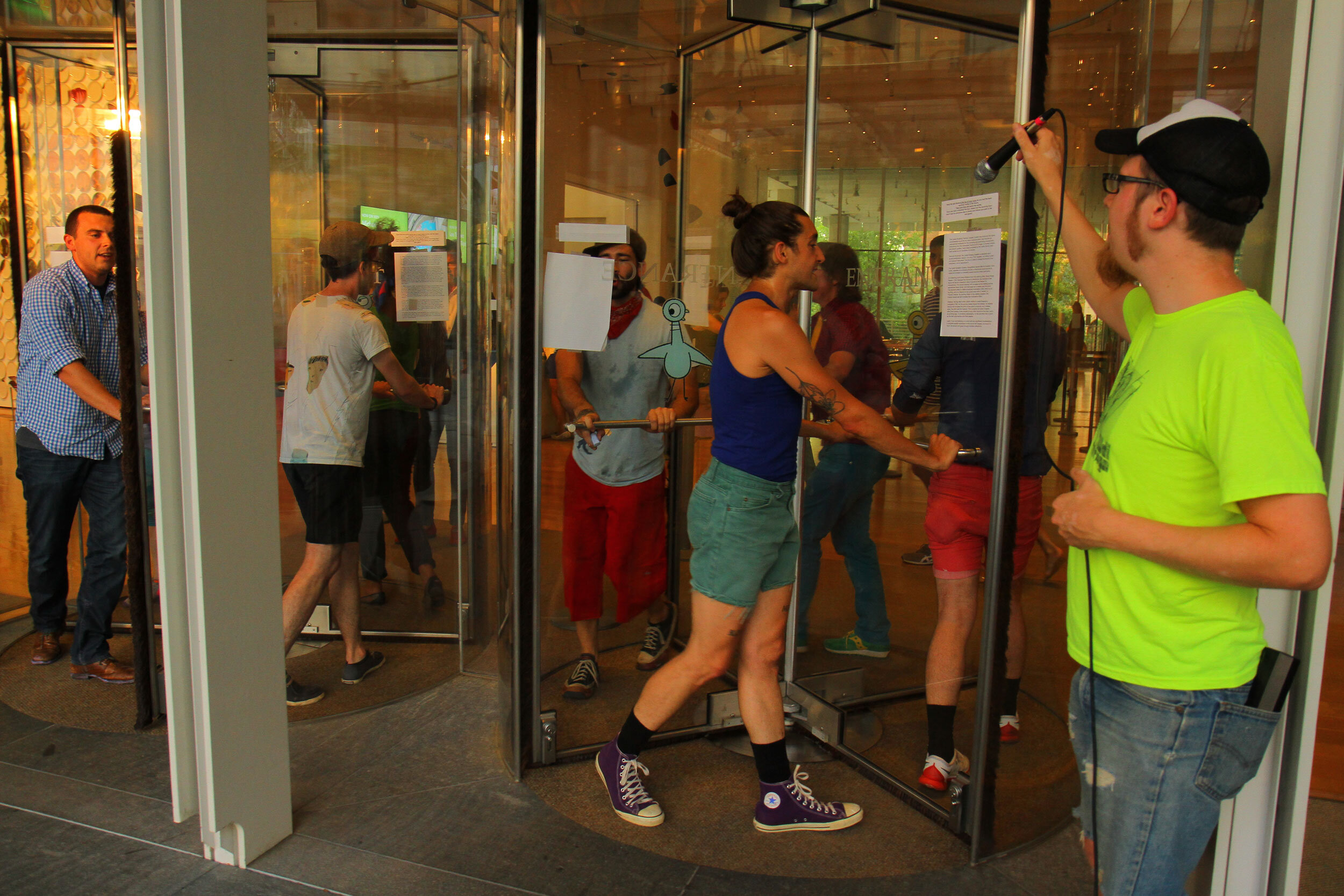 Many performers are walking in circles in 2 revolving glass doors reading text. Another performer is holding up a microphone from outside to catch their voices as they pass.