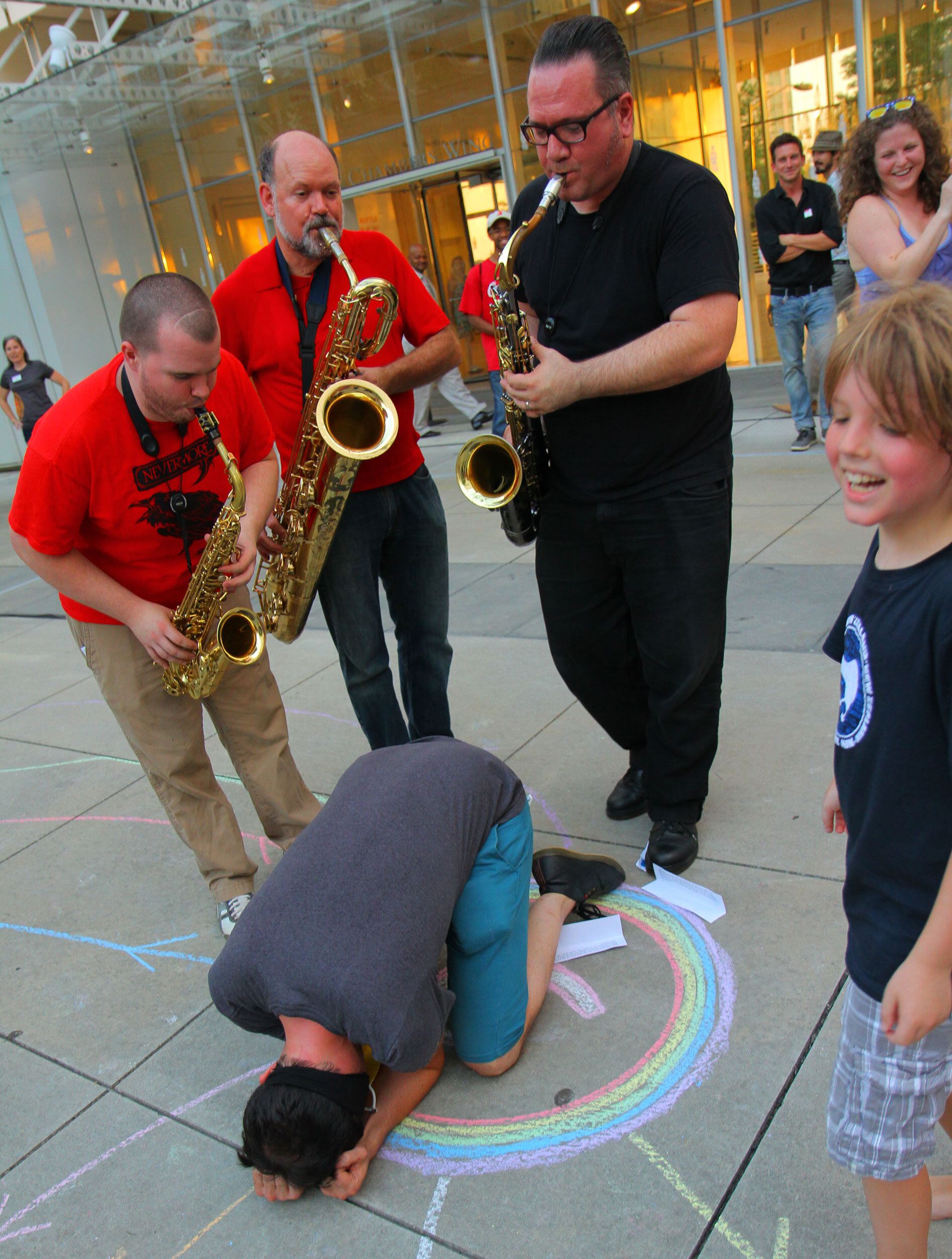 A performer is on the ground on their knees, face down in a gesture of despair. Three musicians stand around them playing saxophones. A boy smiles and laughs nearby.