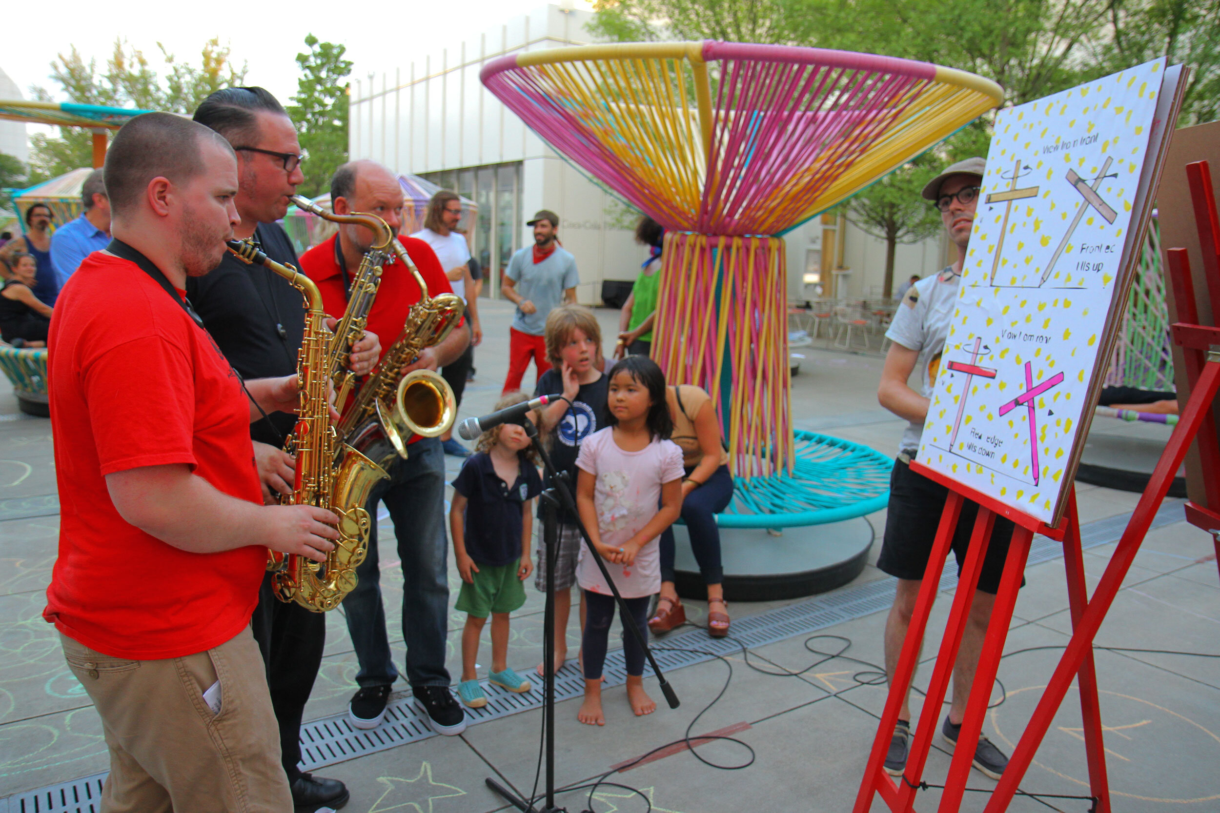 3 musicians with saxophones are looking at a work of art on an easel. A group of children and adults stand by watching them react to the art.