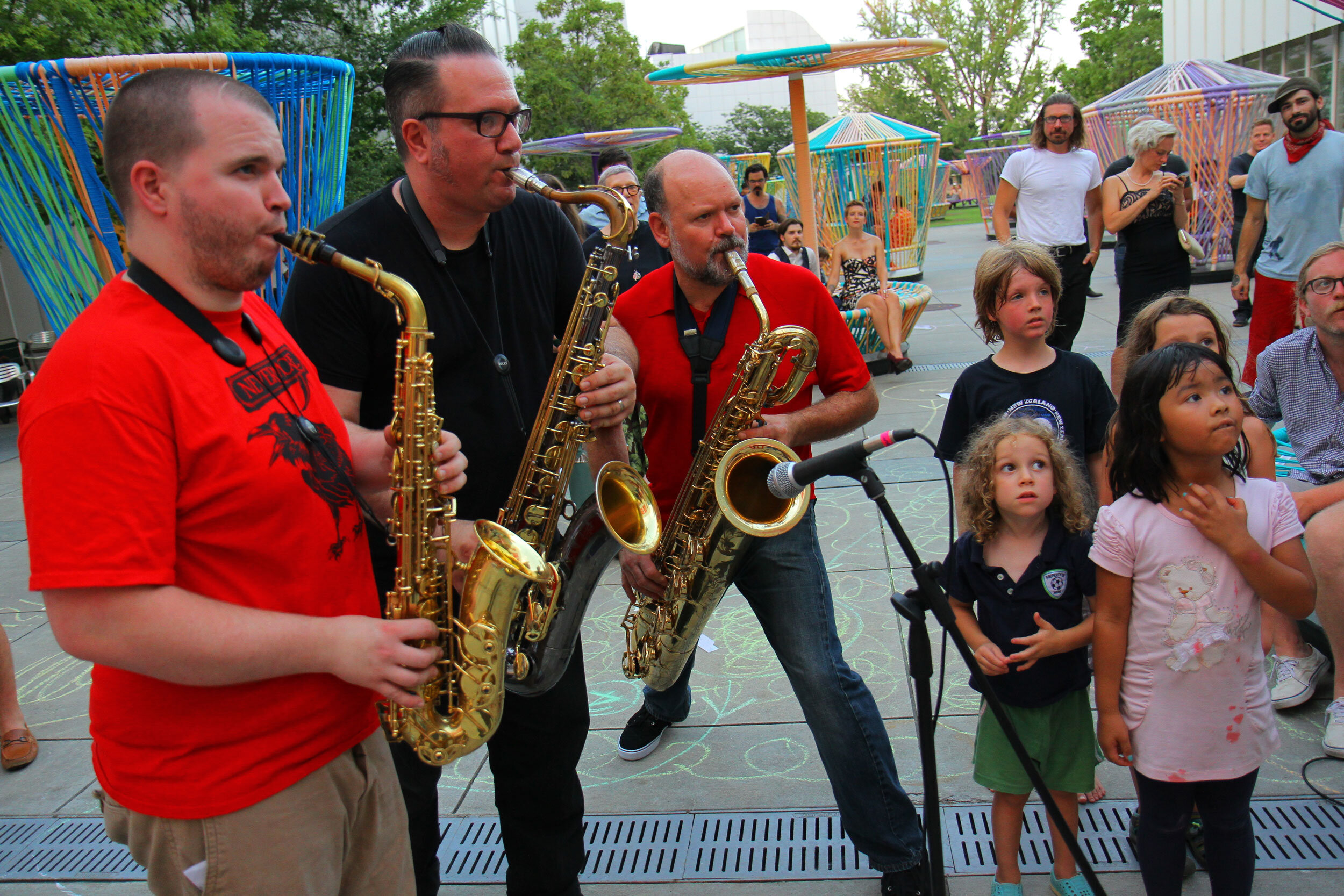 3 musicians with saxophones playing while a group of children watch intently.