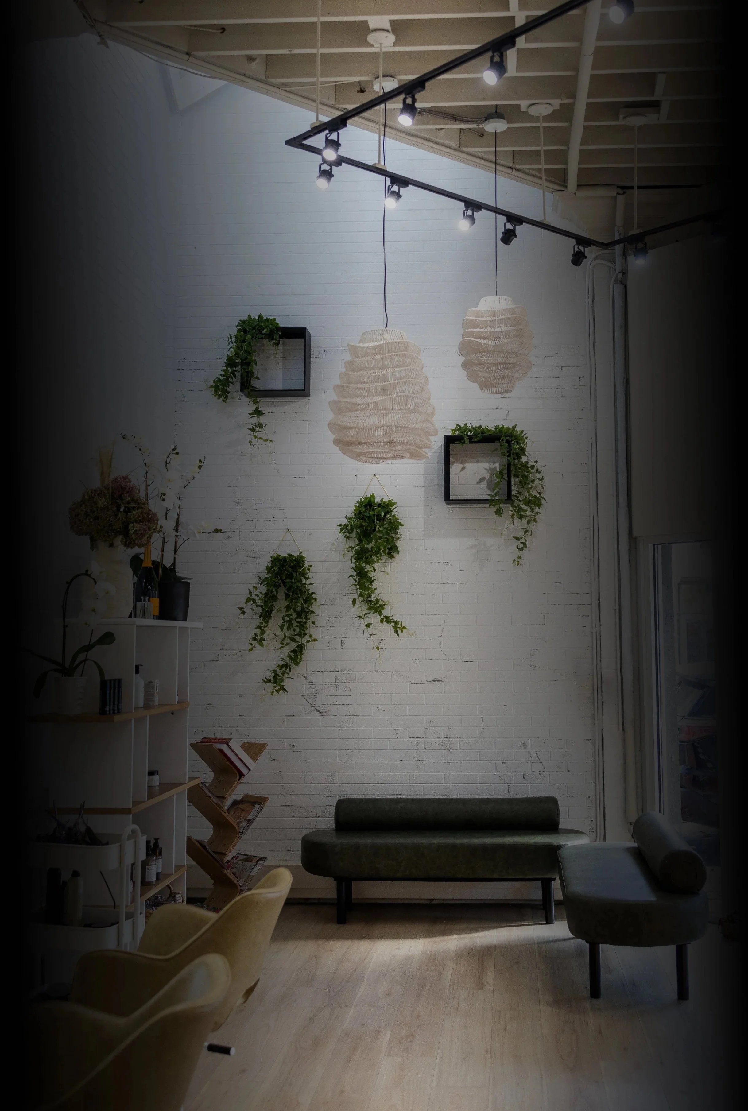 A modern interior room with white brick wall, hanging paper lamp shades, and a bench with a cylindrical cushion. There are shelves with plants and decor items, and ceiling track lighting.
