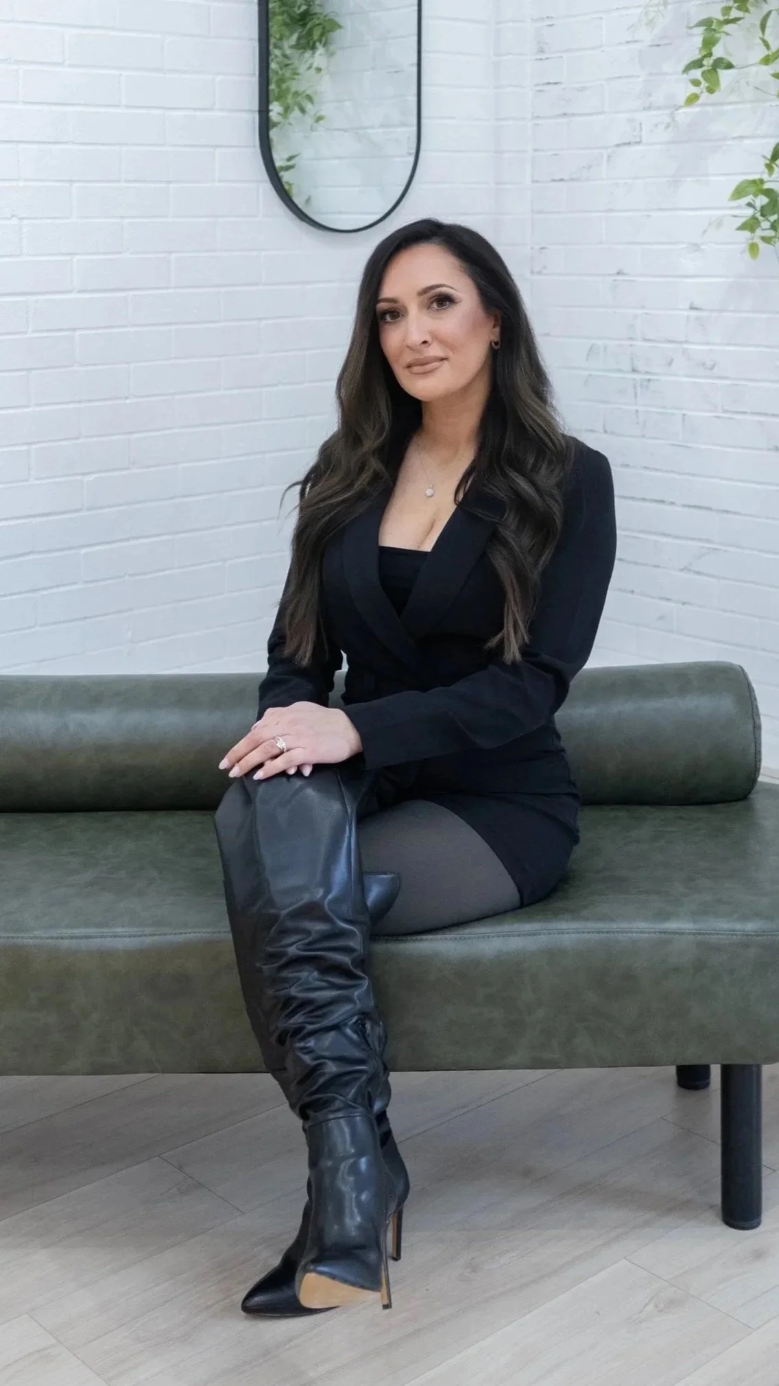 A woman with long dark hair, wearing a black blazer, sitting on a green bench in a modern interior with a white brick wall and a mirror behind her.
