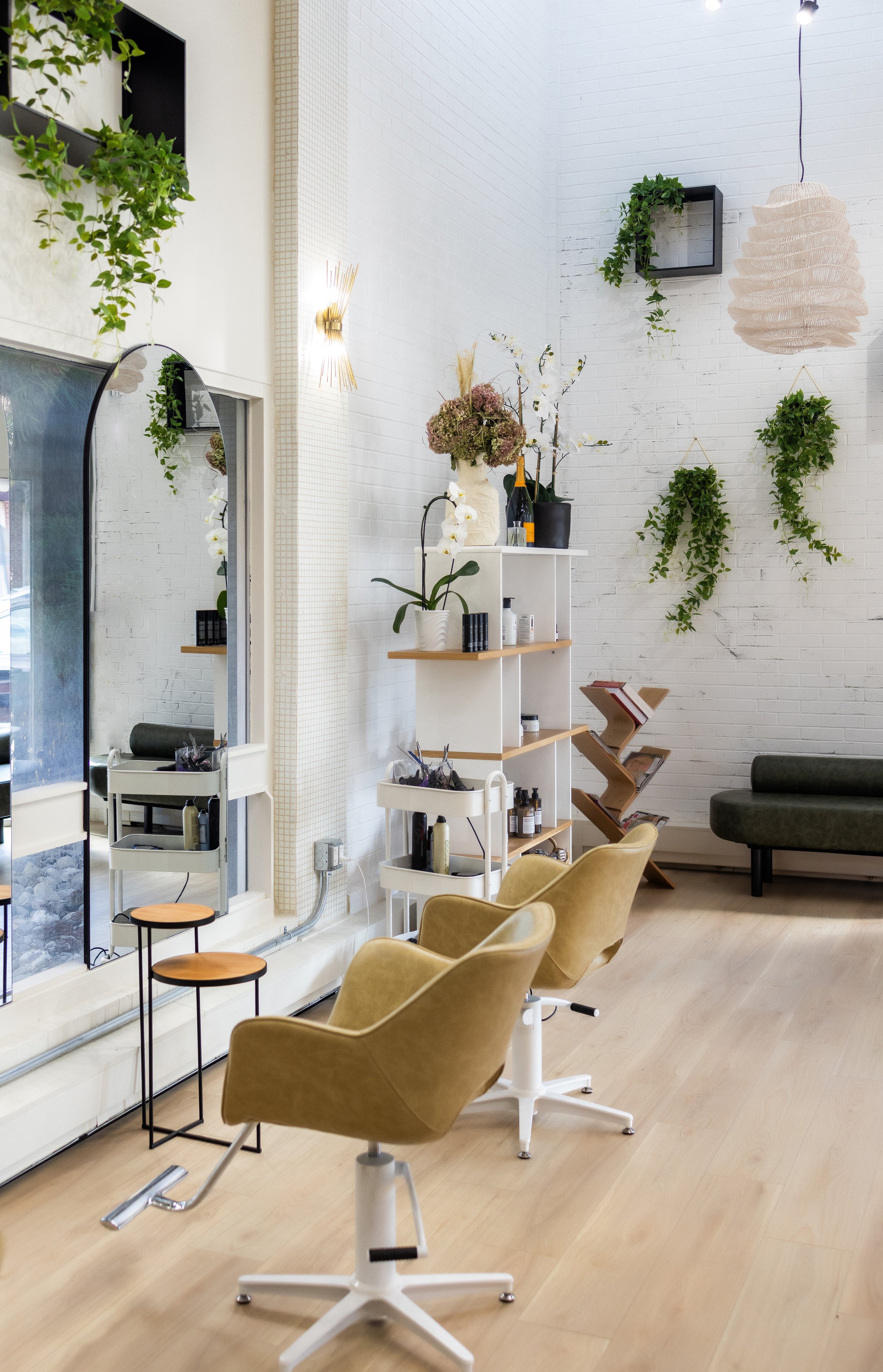 Interior view of a beauty salon or hair studio featuring chairs, styling stations, and decorative plants on white walls, with a large mirror and wooden flooring.