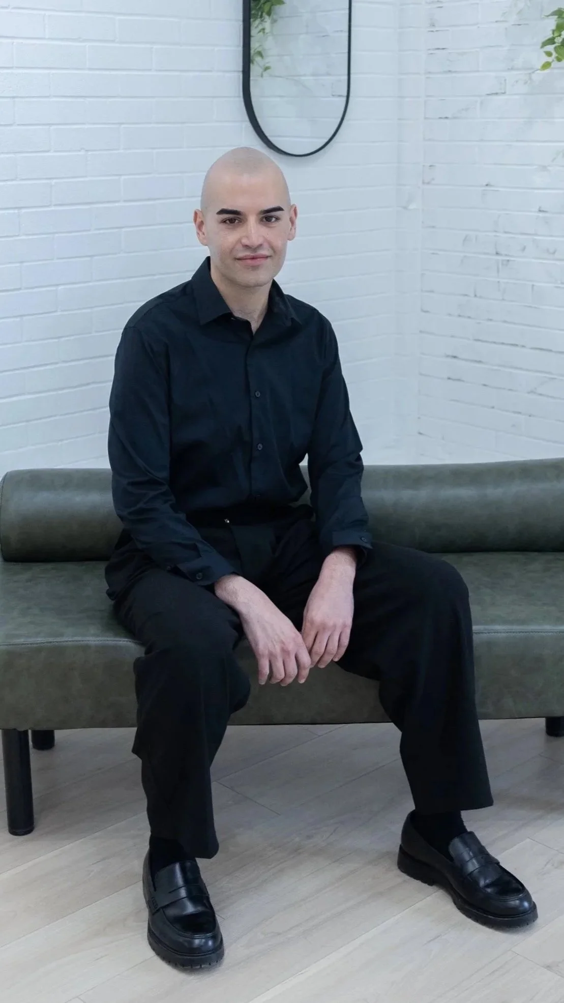 A man with a shaved head wearing a black shirt and black pants sitting on a green bench against a white brick wall.