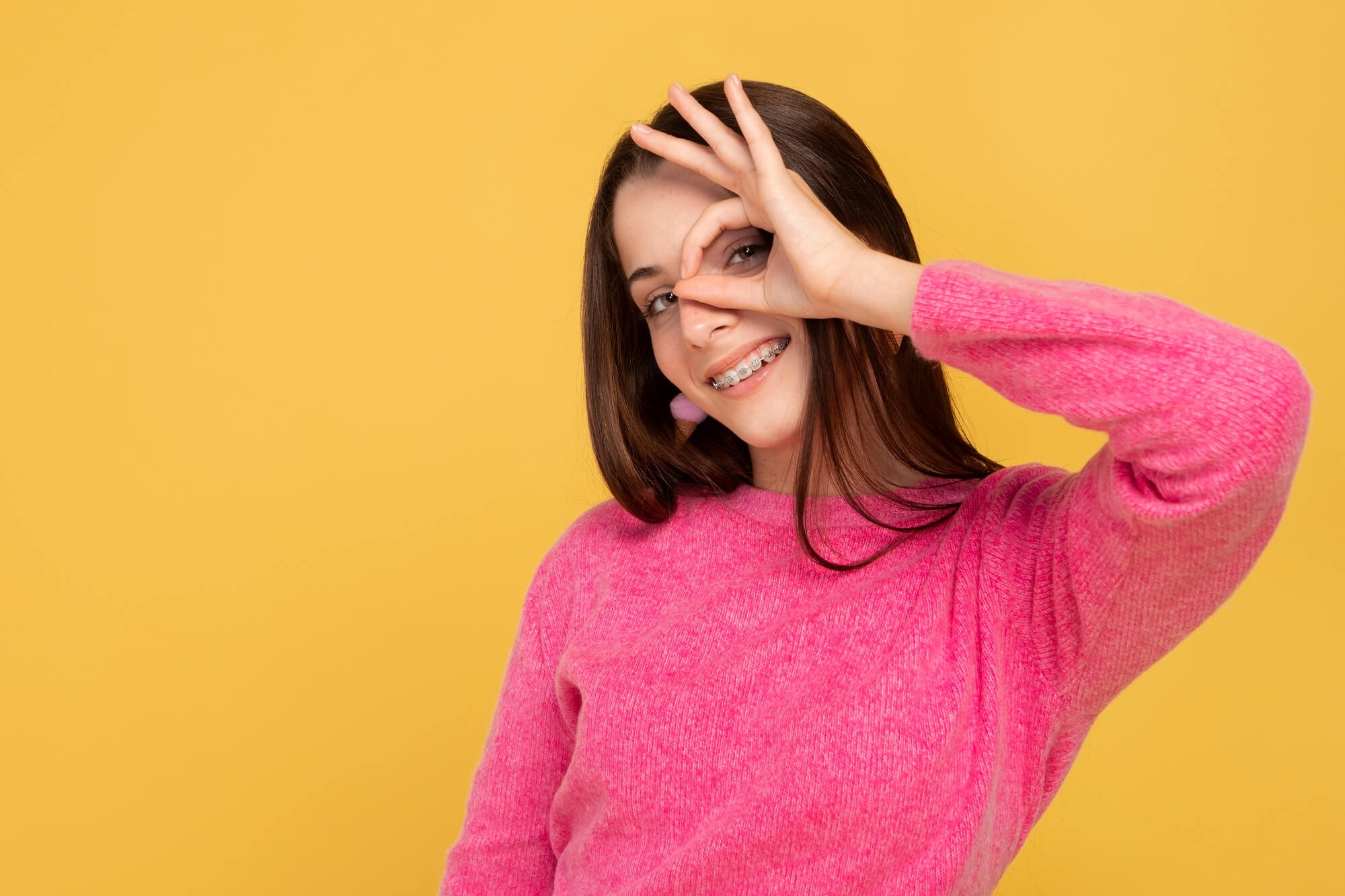 Young woman with brown hair smiling and making an 'okay' sign around her eye with her fingers, wearing braces and a pink sweater against a yellow background.