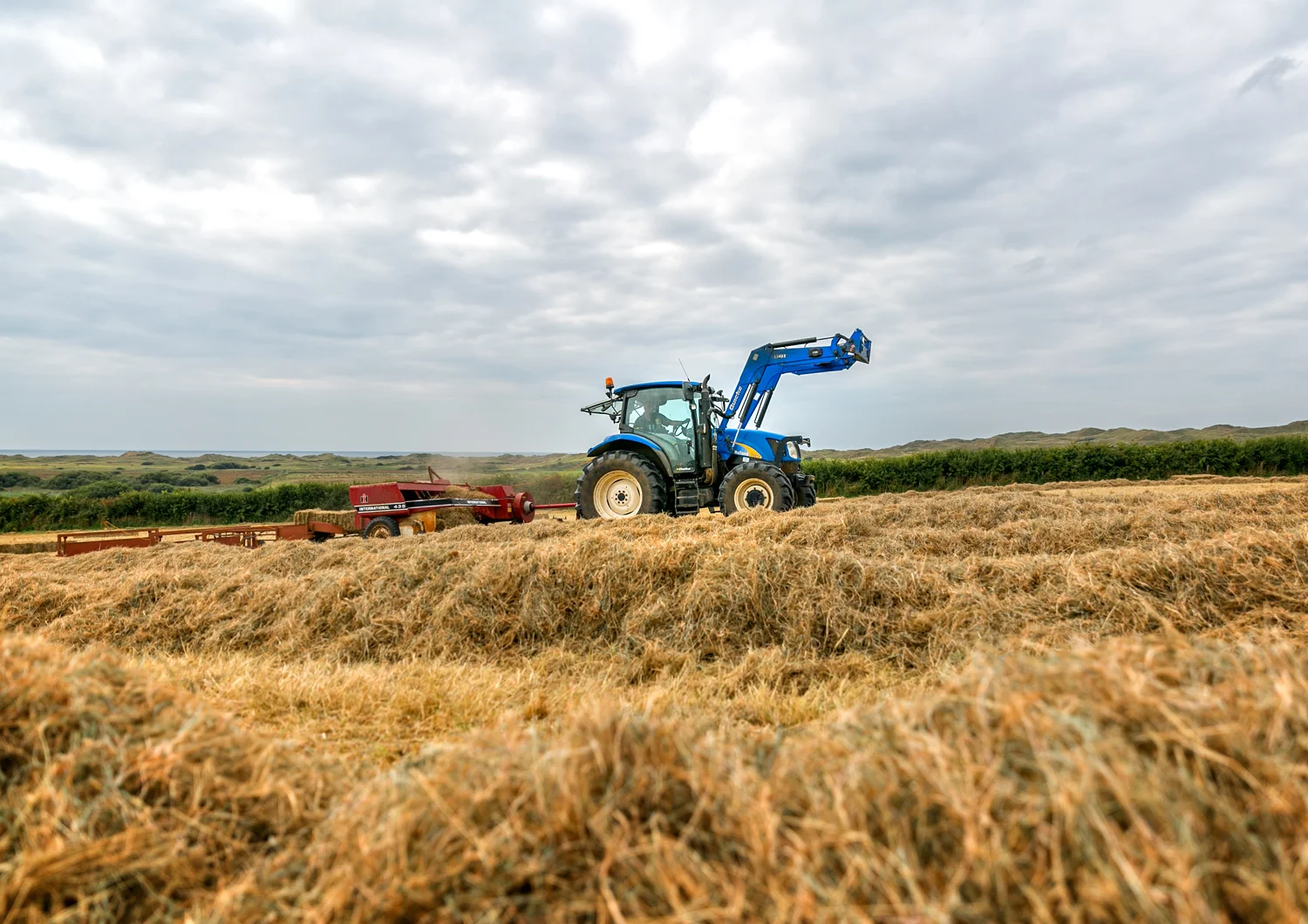 Blue tractor moving through a field of dried crops under a cloudy sky with a distant landscape of green trees and rolling hills.