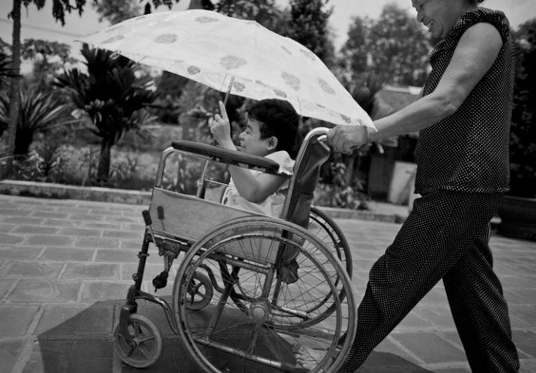 Thao is being pushed in her wheelchair by her mother. The family wheels Thao out to the separate structure that houses Thao’s library along with her father’s pig feed and fertilizer. There Thao will sit in her library for a majority of the day excepting local children from the village.