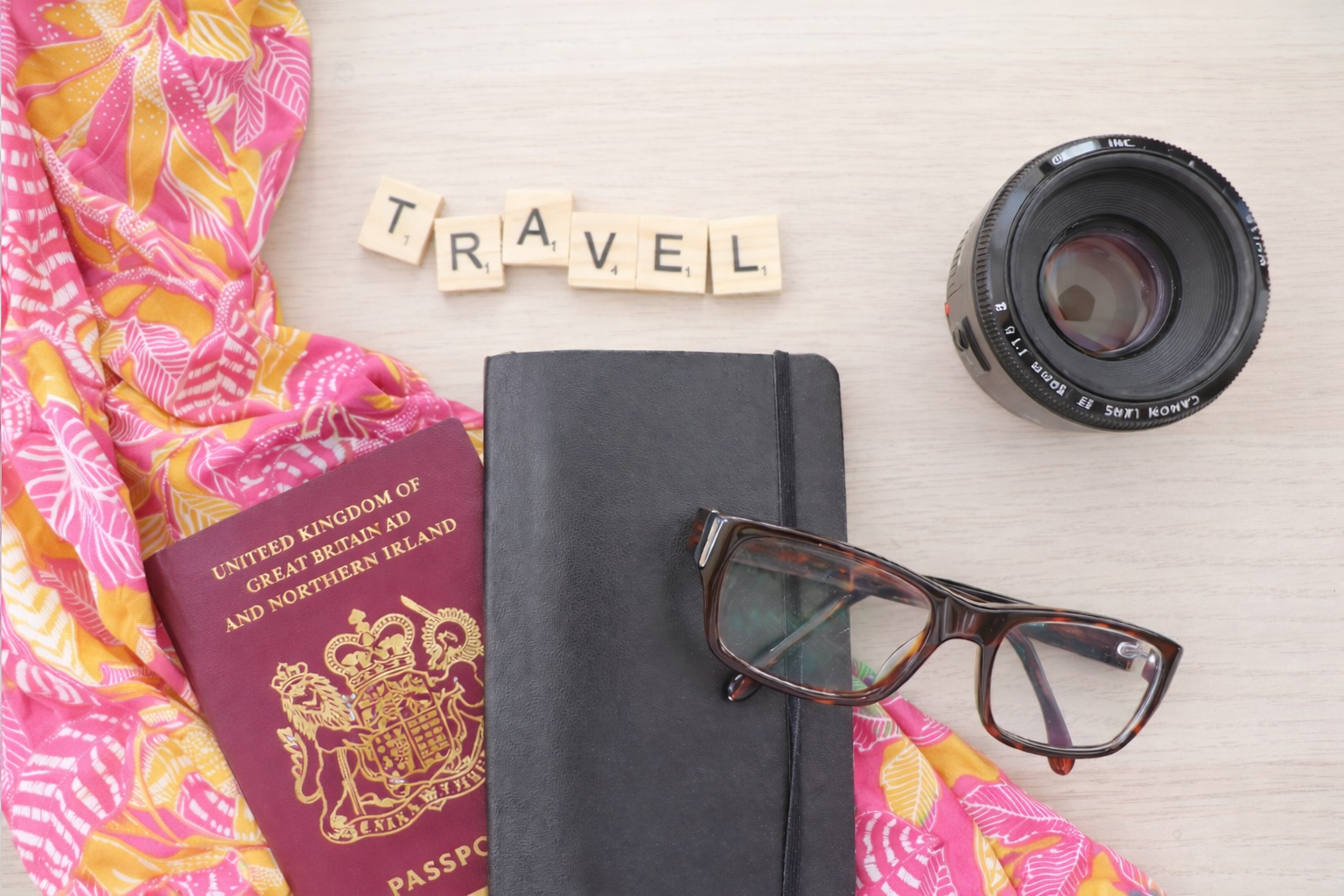 Flat lay of travel essentials including a UK passport, black notebook with glasses, camera lens, and pink and yellow scarf, with Scrabble tiles spelling travel on a light wooden surface