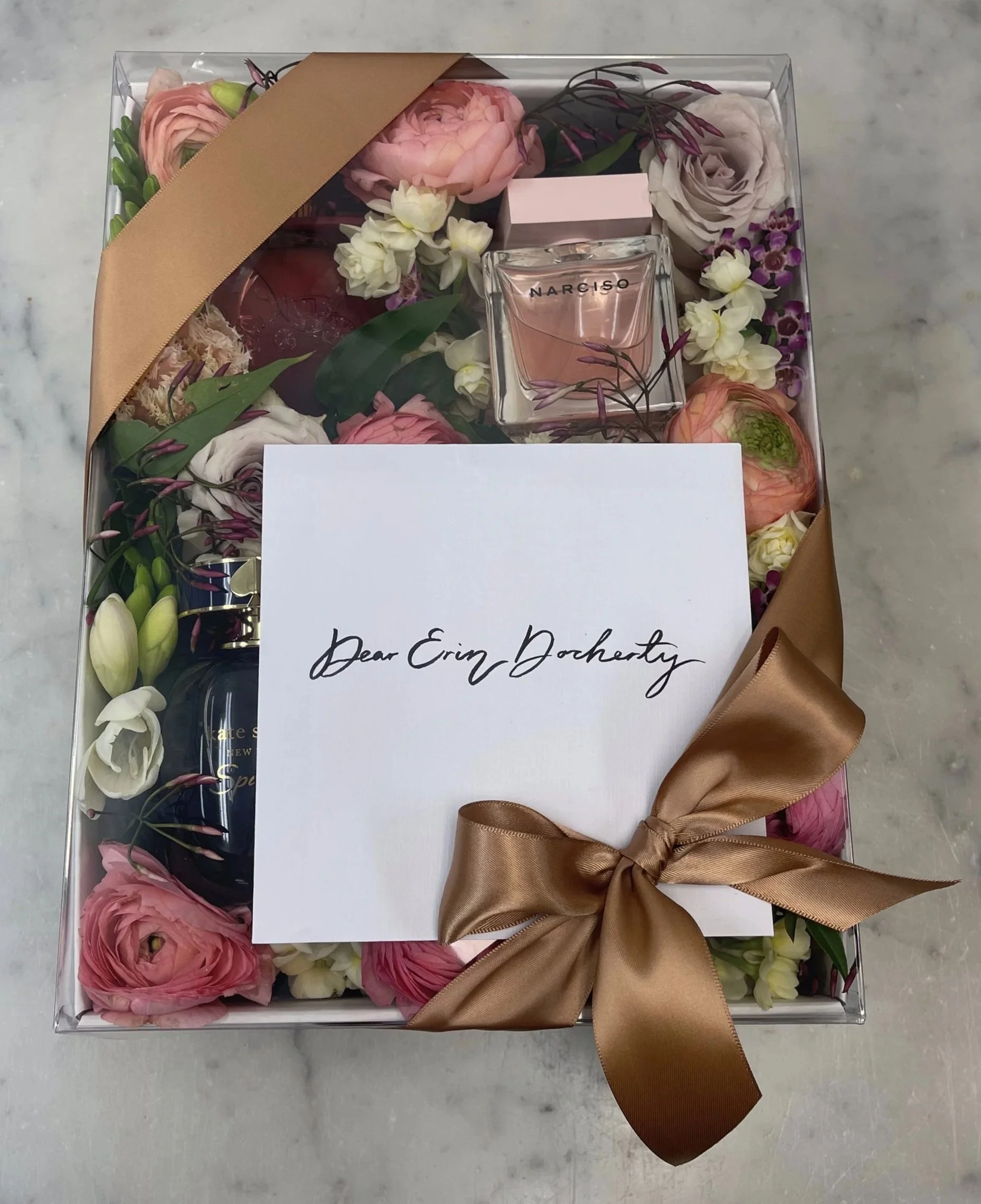 A gift box with pink and white flowers, a perfume bottle labeled Narciso, a small pink box, and assorted beauty and skincare products inside, with a white note that says 'Dear Erin Doherty' and a large brown ribbon tied around the box.
